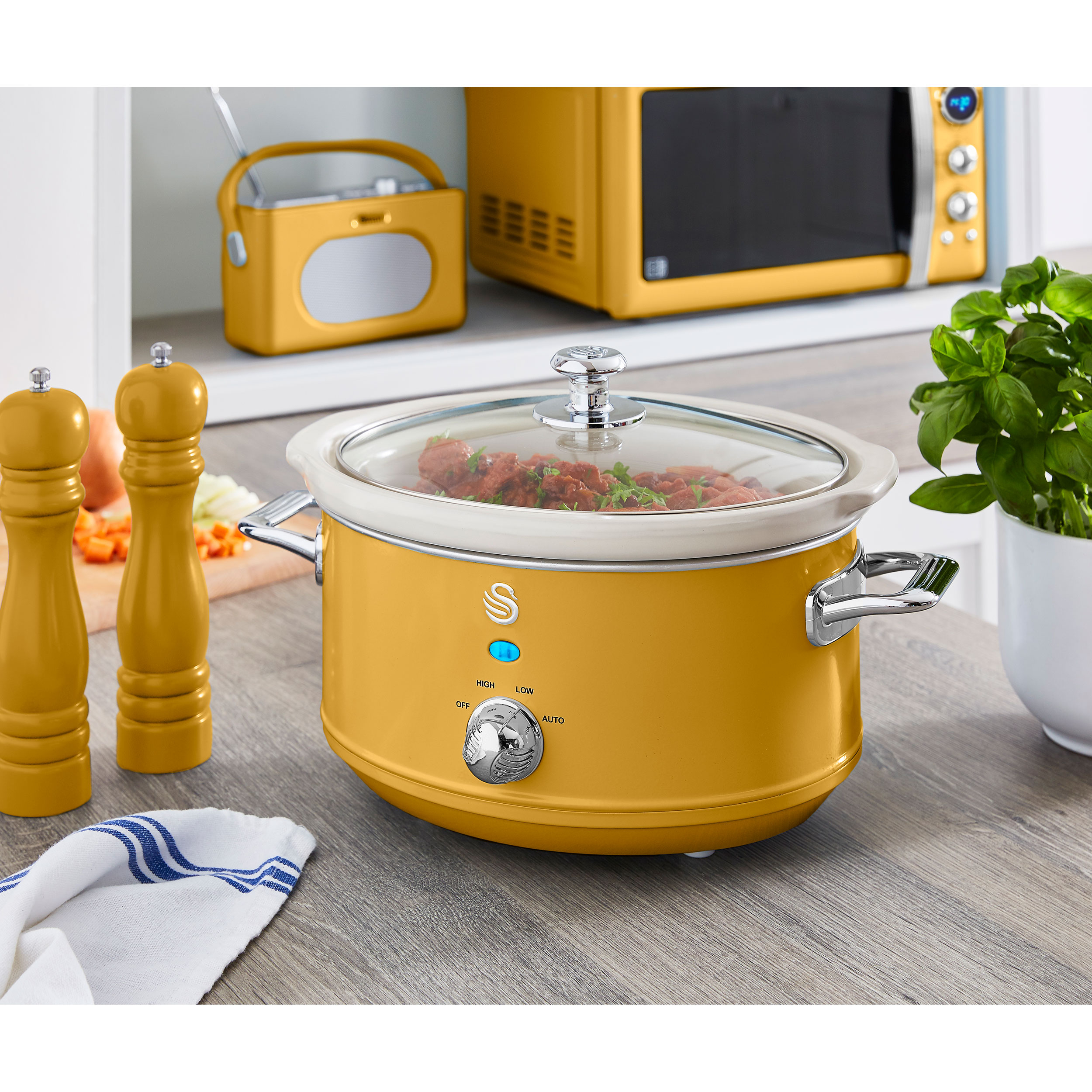 Olla de cocción lenta amarilla con tapa de cristal, sobre una mesa con ingredientes y un microondas detrás.