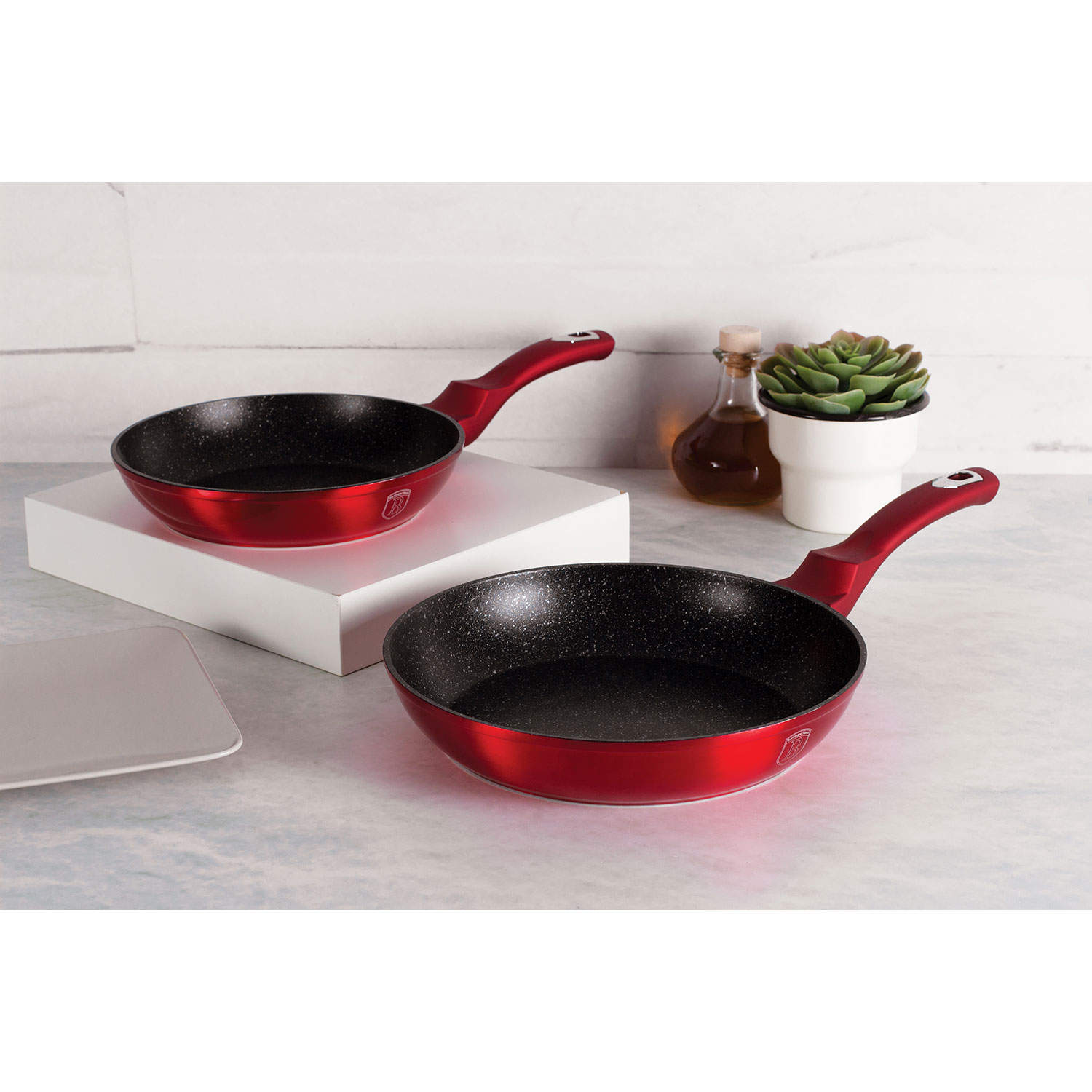Dos sartenes rojas con interiores moteados negros y mangos rojos, sobre una superficie blanca.
