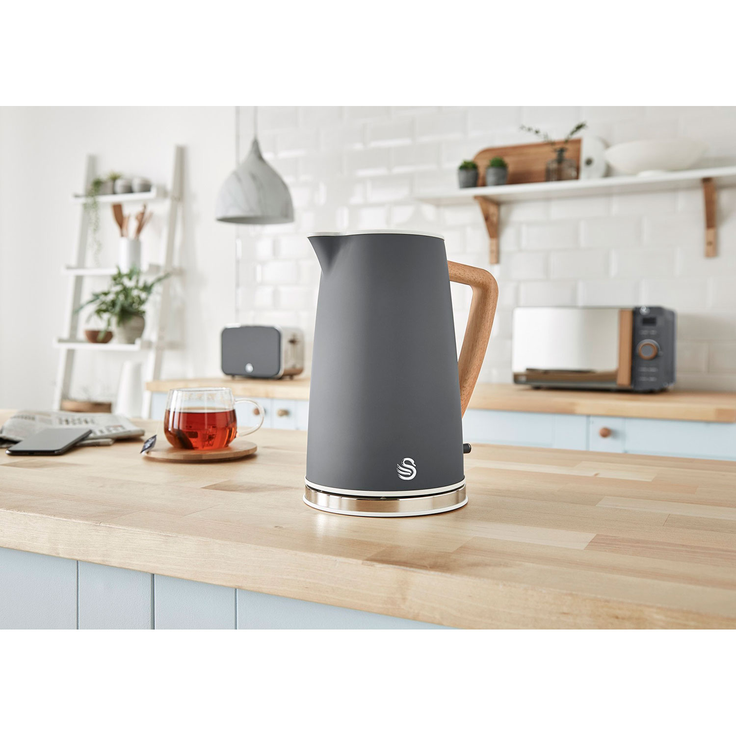 Ein grauer Wasserkocher mit Holzgriff, eine Teetasse und ein Toaster auf einer Holztheke.