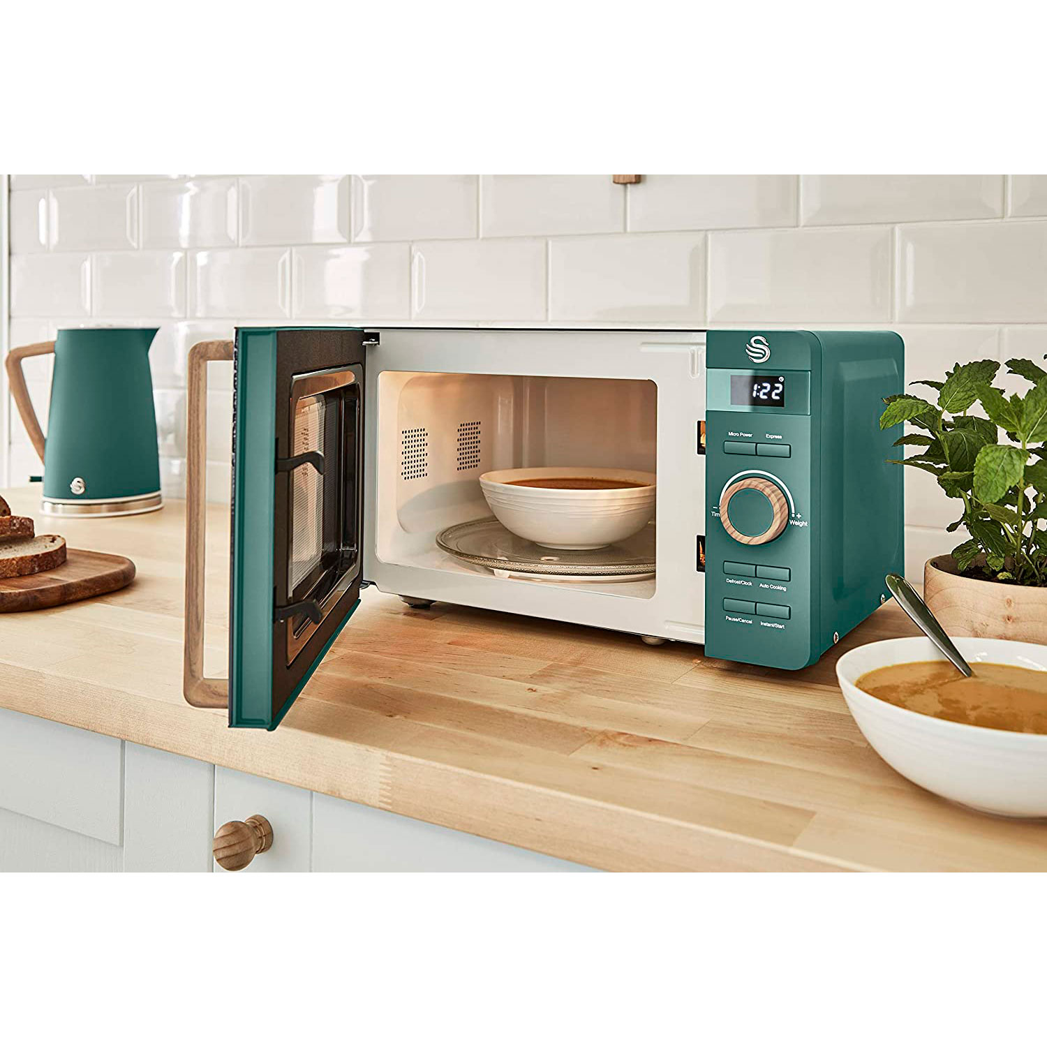 Microondas turquesa con puerta abierta, cuenco dentro. Asa de madera, tetera turquesa y comida en tabla de madera.