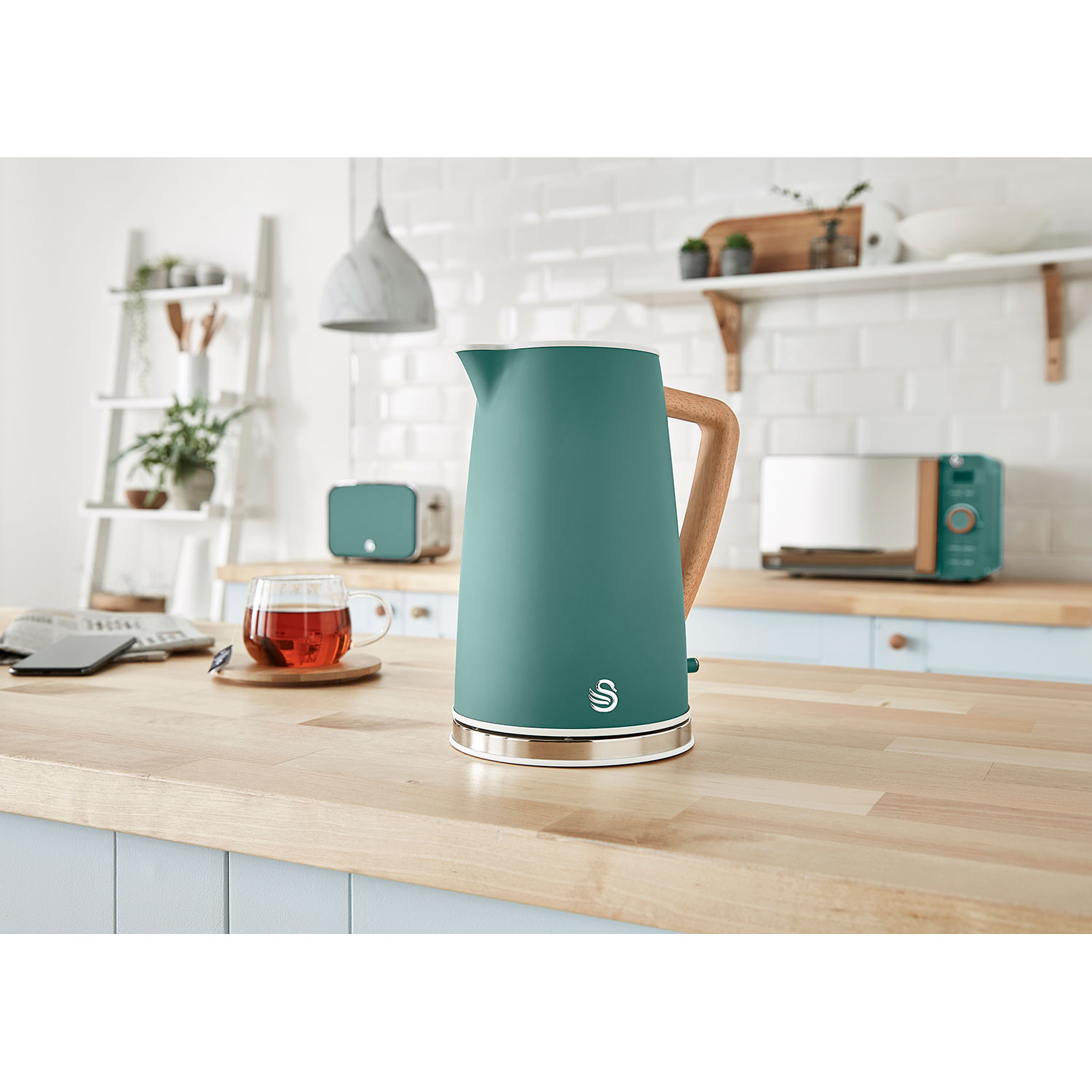 Grüner Wasserkocher mit Holzgriff, Teeservice und Küchengeräte auf Holzoberfläche.