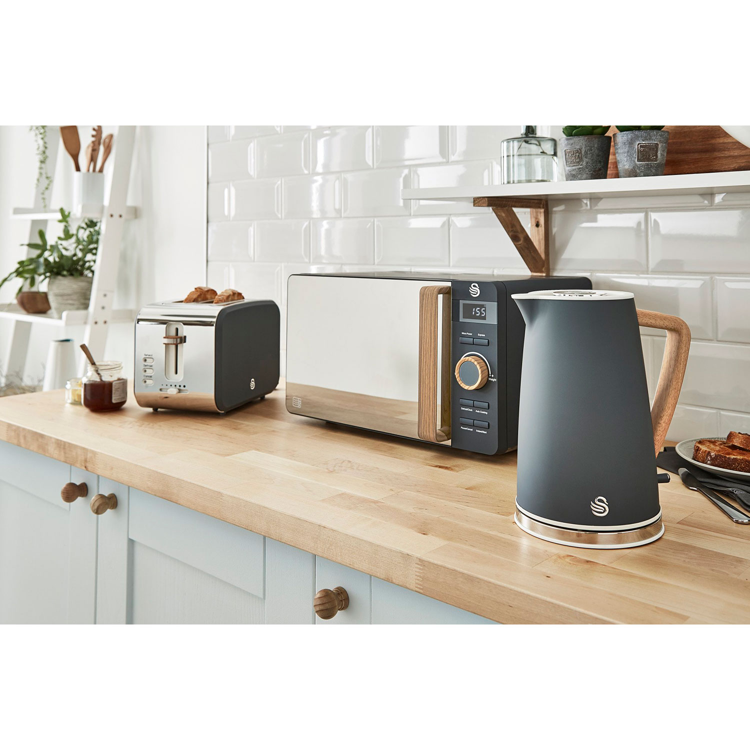 Küchengeräte auf einer Holztheke: Mikrowelle, Toaster und Wasserkocher. Weißer Fliesen-Hintergrund.