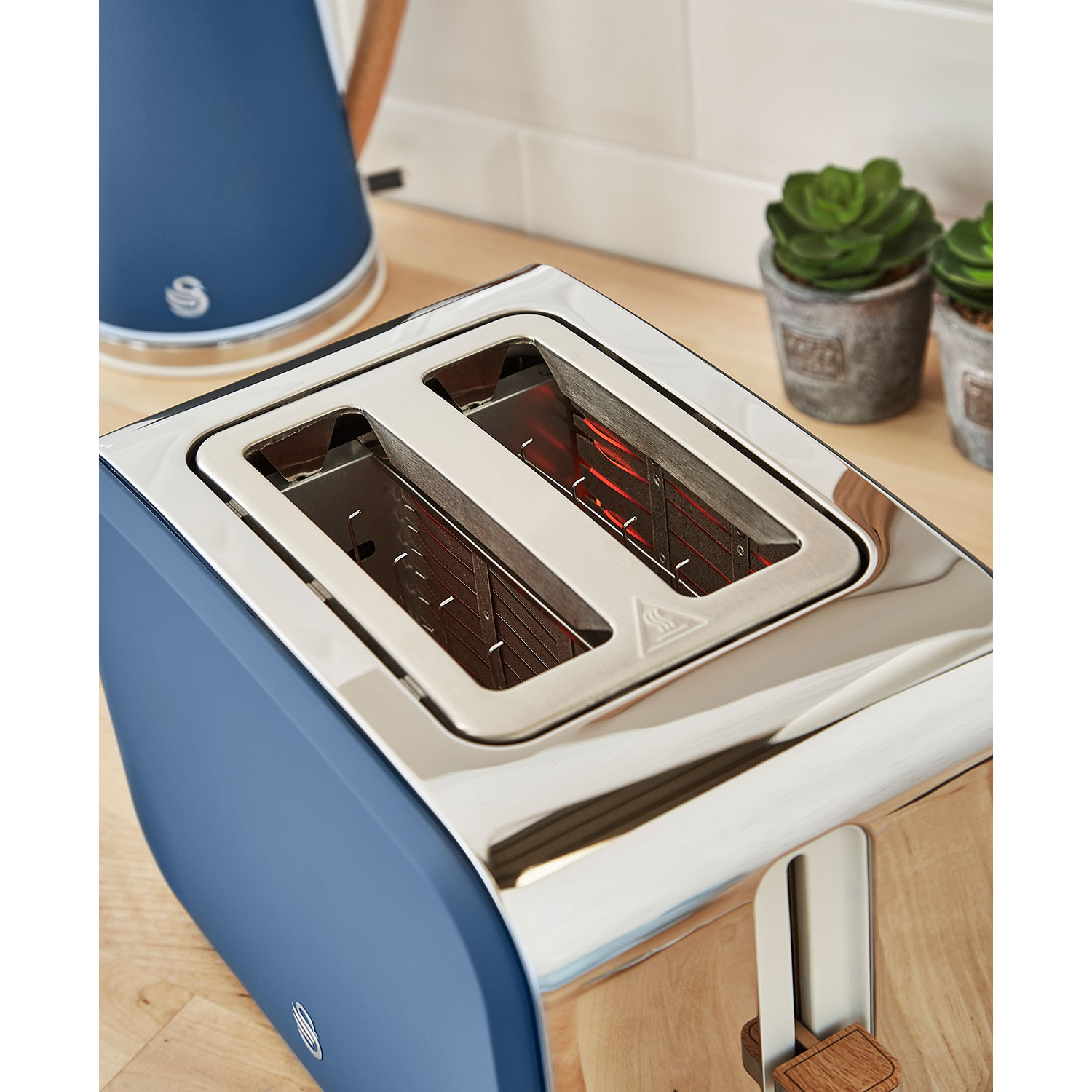 Blauer Toaster mit zwei Schlitzen, mit Holzgriff, in der Nähe eines Wasserkochers und Pflanzen.