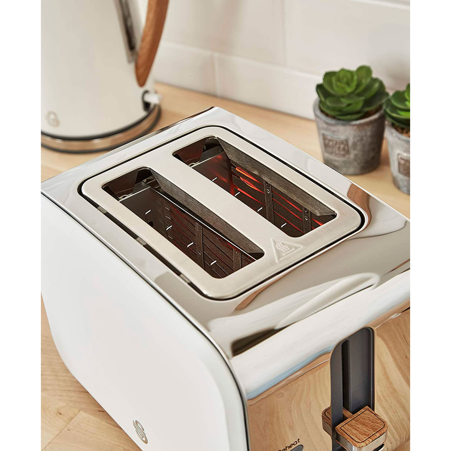 Weißer und silberner Toaster mit zwei Schlitzen, ein Wasserkocher und Pflanzen im Hintergrund.