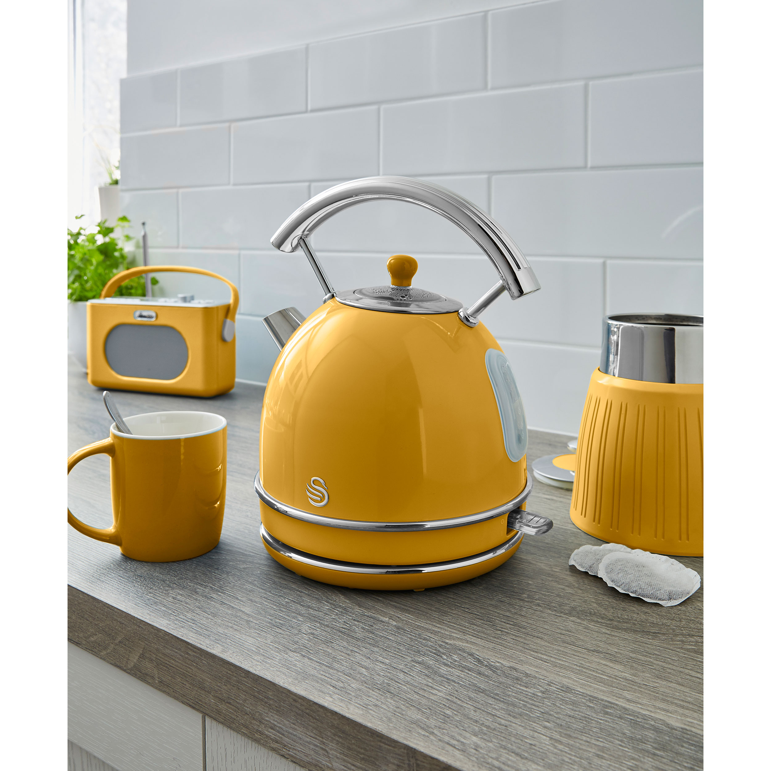 Hervidor y taza amarillos de estilo retro en una encimera, junto a una radio amarilla e infusor de té.
