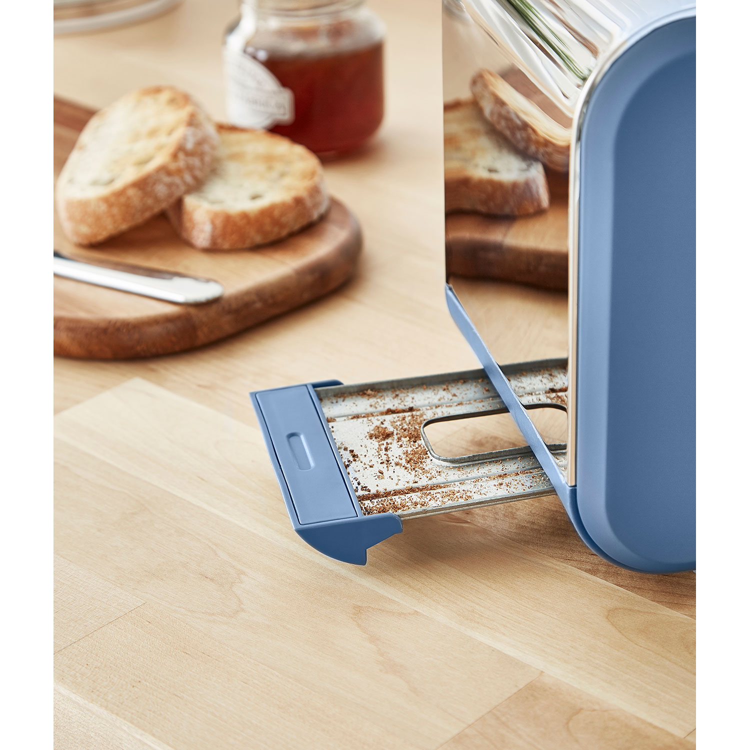 Ein Toaster mit herausgezogener Krümelschublade, die Krümel zeigt. Brot und Marmelade liegen auf einem Holzbrett.