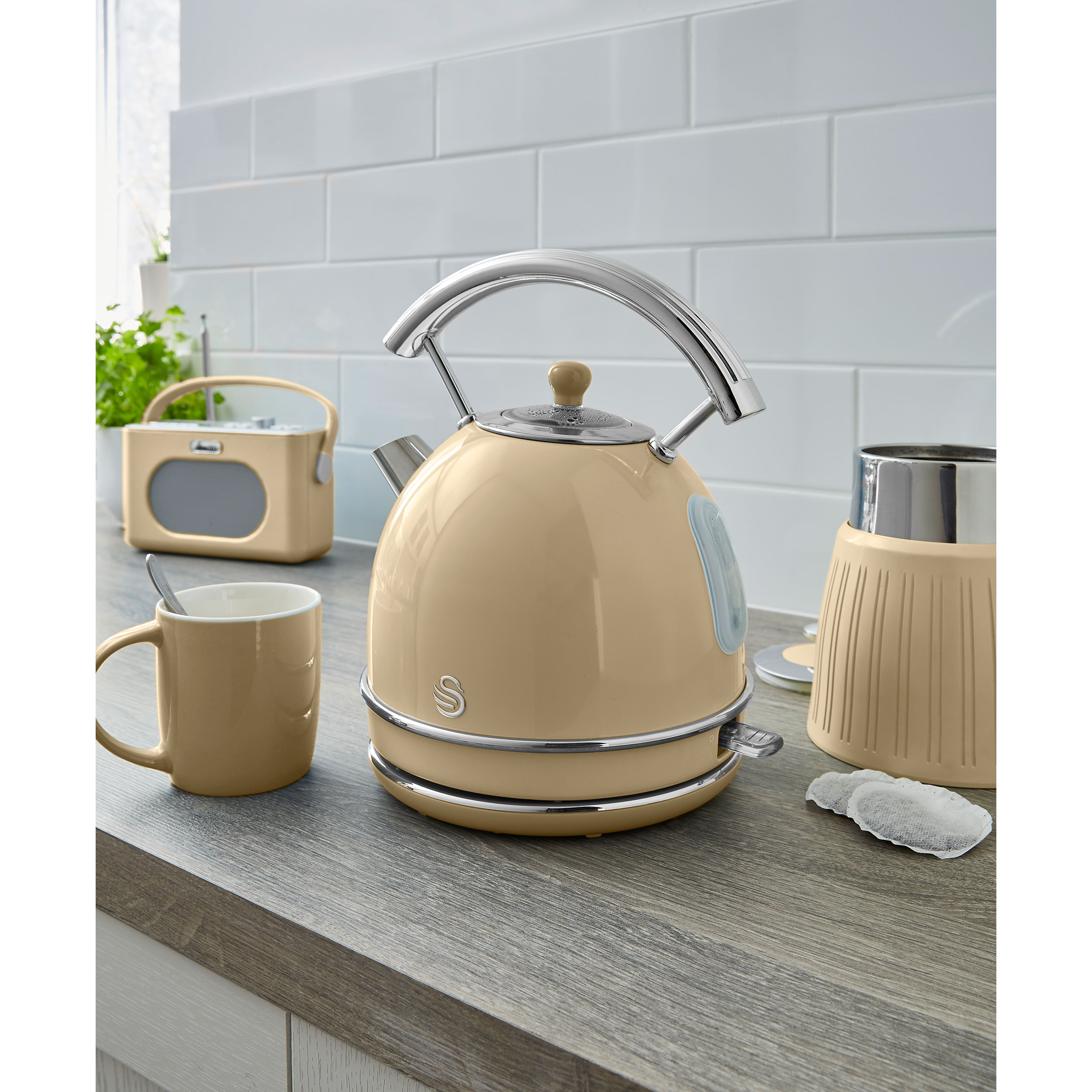 Hervidor beige, taza y accesorios en mostrador de madera, pared de azulejos blancos detrás.