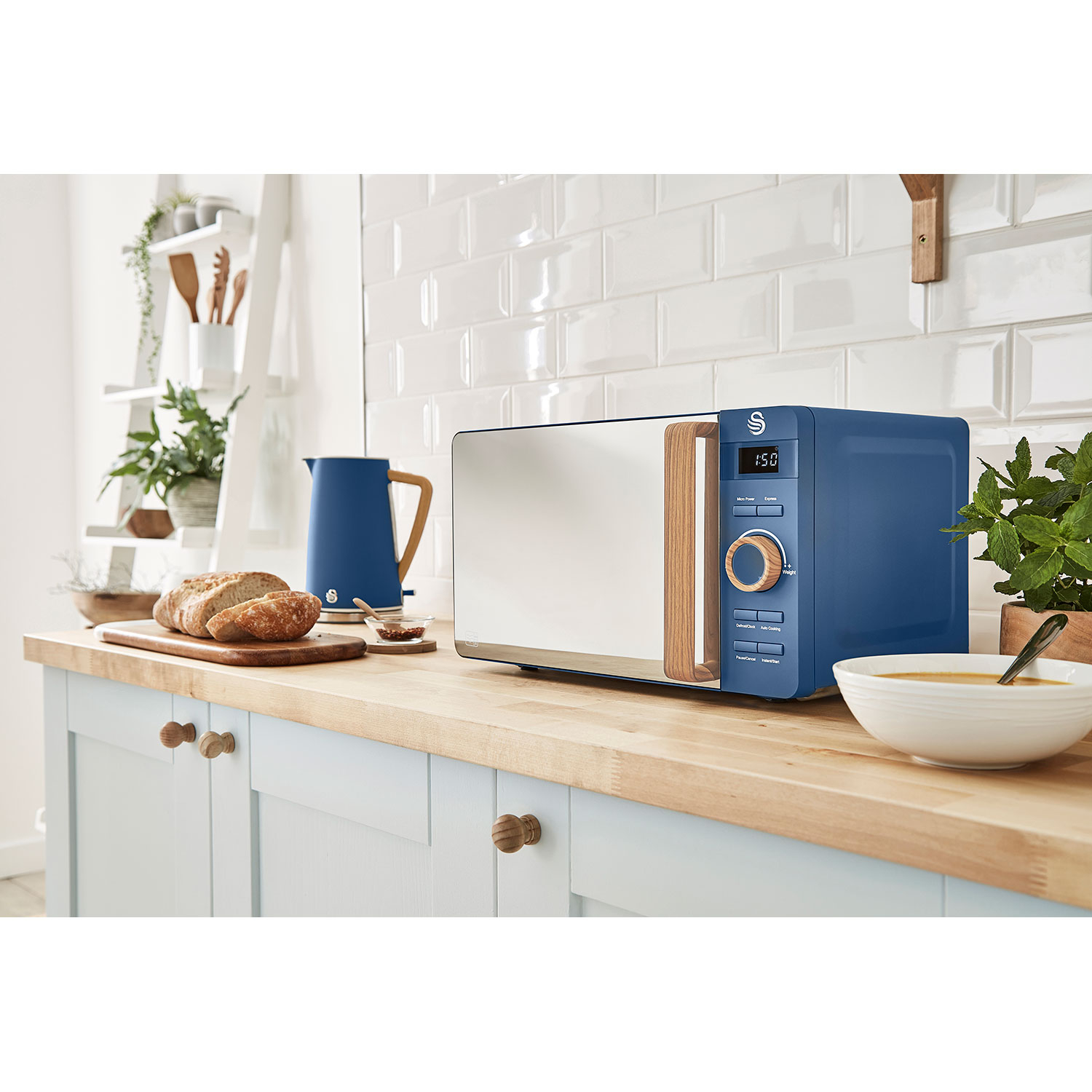 Un horno microondas azul sobre una encimera de madera en un entorno de cocina. Hay una tetera azul y pan.