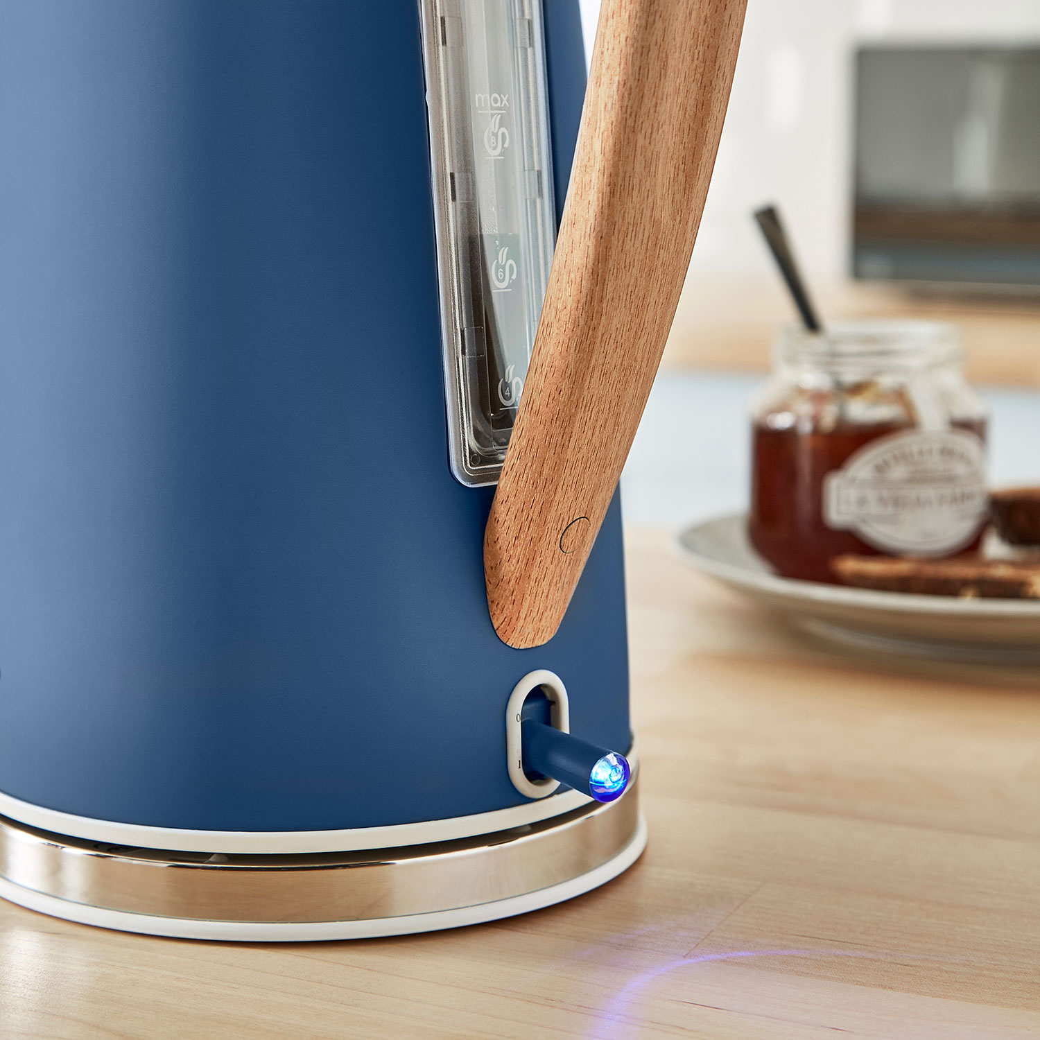 Blauer Wasserkocher mit Holzgriff. Ein Licht ist an. Ein Glas Marmelade und Brot sind daneben.