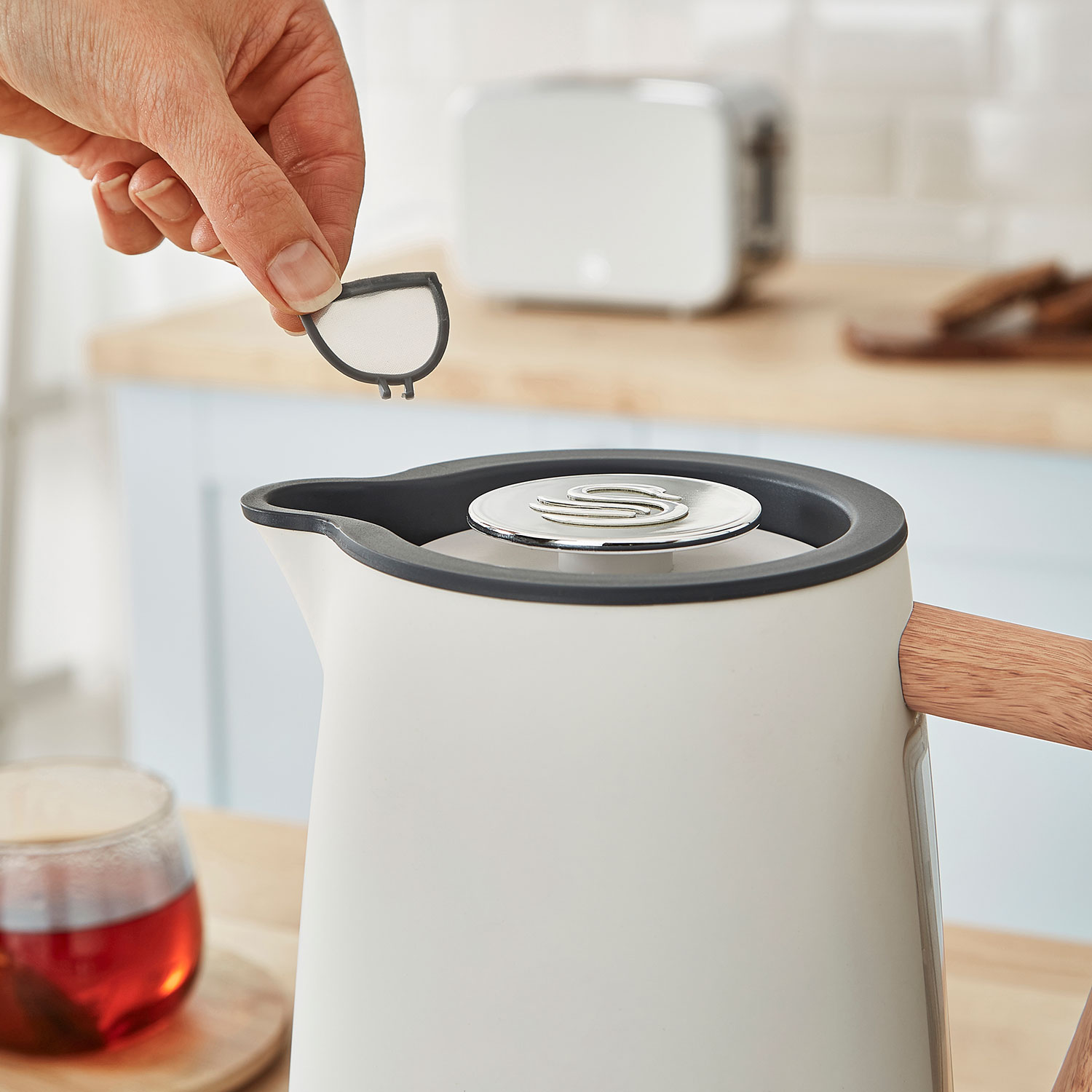 Dłoń trzymająca filtr nad białym czajnikiem z drewnianymi akcentami.