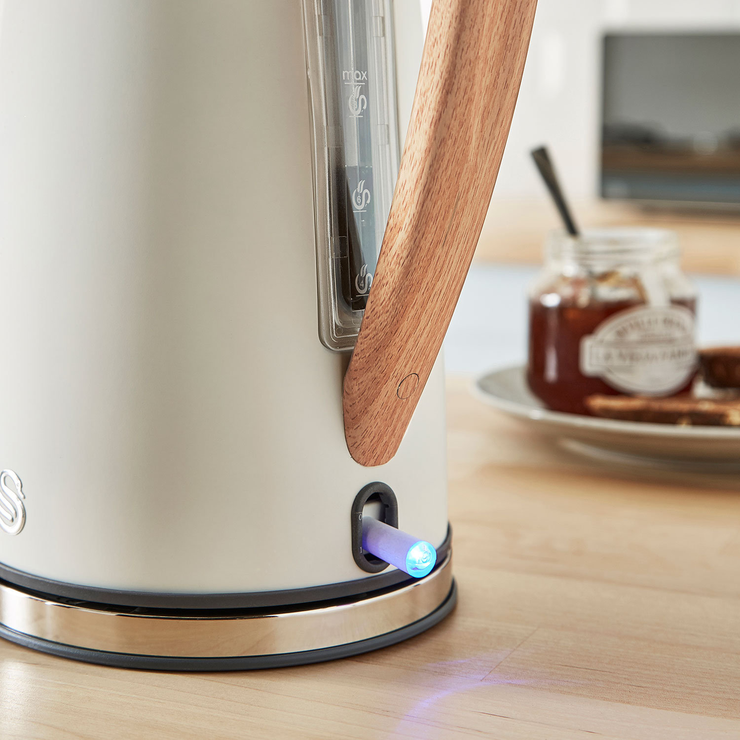 Biały czajnik z drewnianym uchwytem. Niebieskie światło u podstawy. Słoik i talerz w tle.