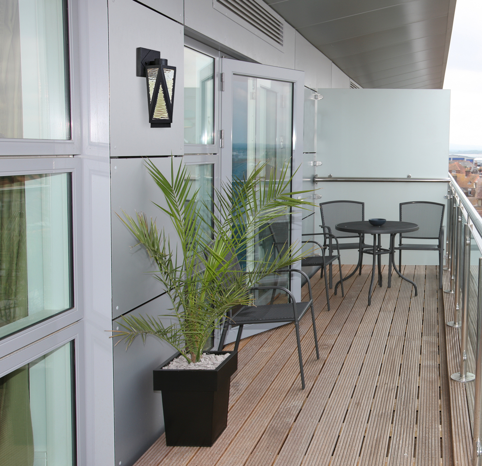 Balkon mit Holzboden, Metallstühlen und -tisch. Eine hohe Topfpflanze und eine an der Wand befestigte schwarze Leuchte.