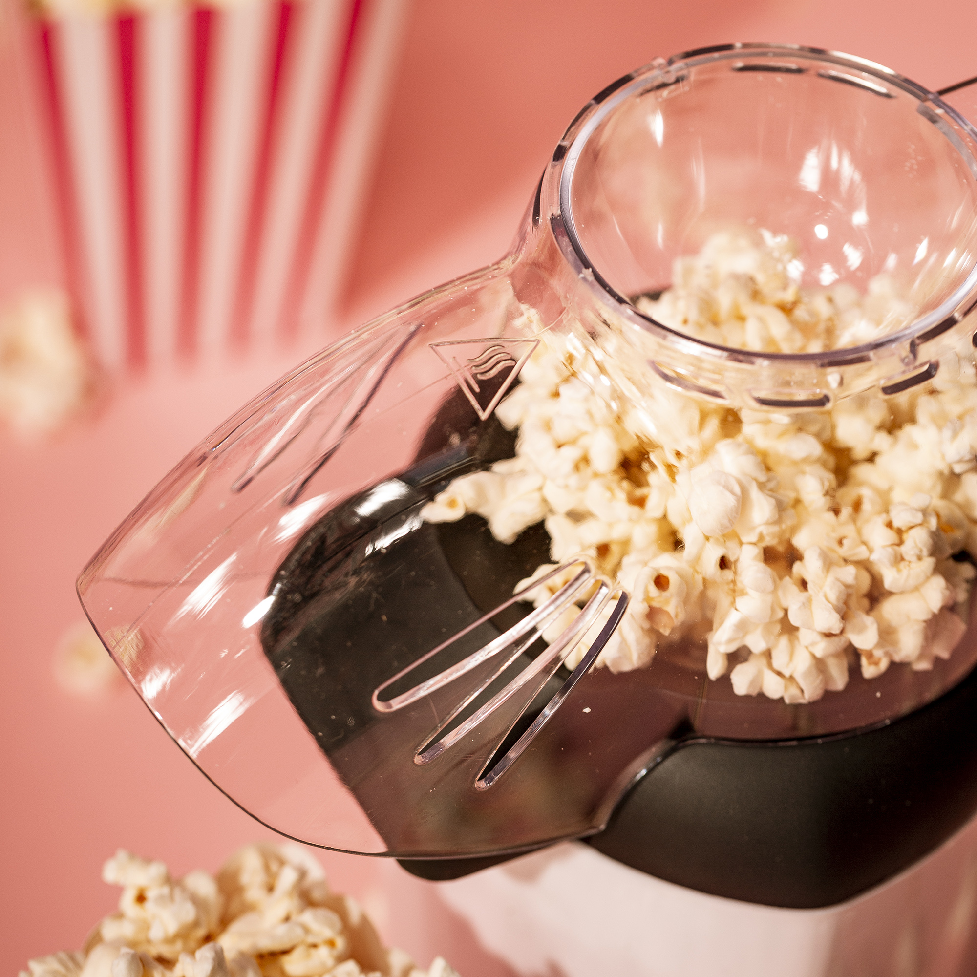 Máquina de palomitas con palomitas dentro, derramándose sobre una superficie rosa con fondo a rayas.