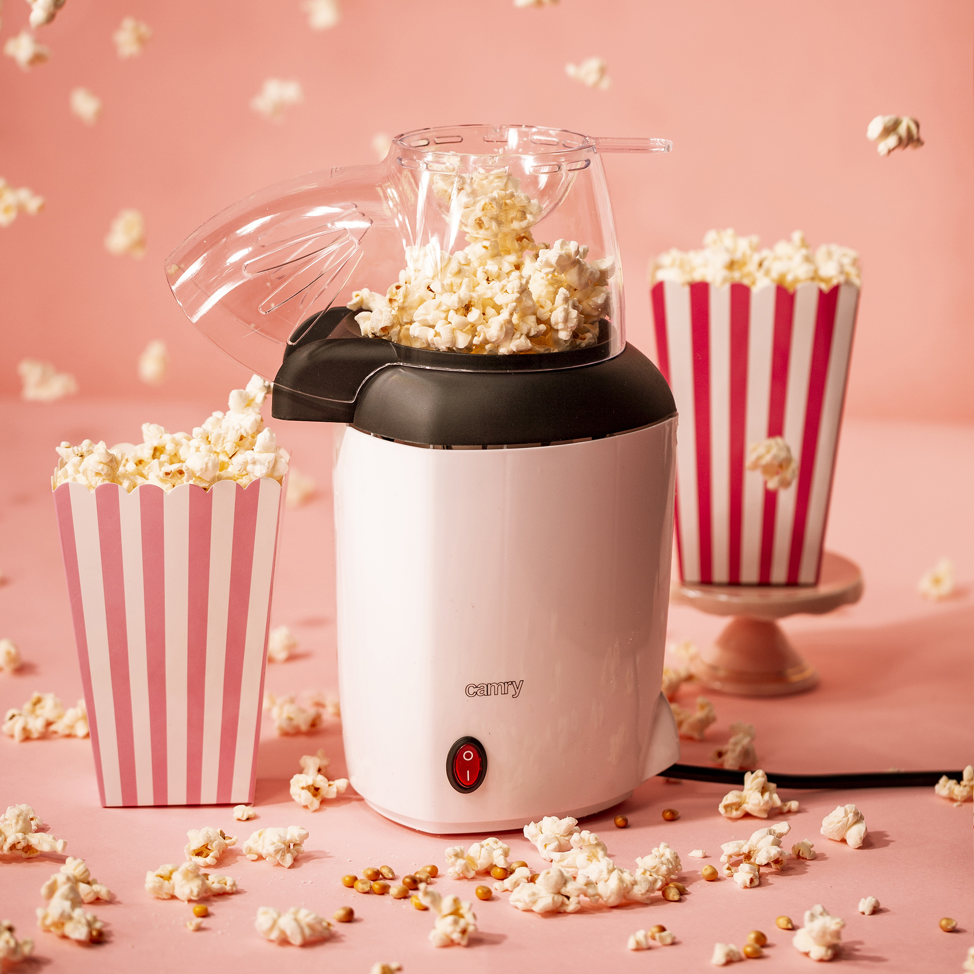 Una máquina de palomitas blanca con palomitas y dos cajas de palomitas a rayas rosas sobre fondo rosa.