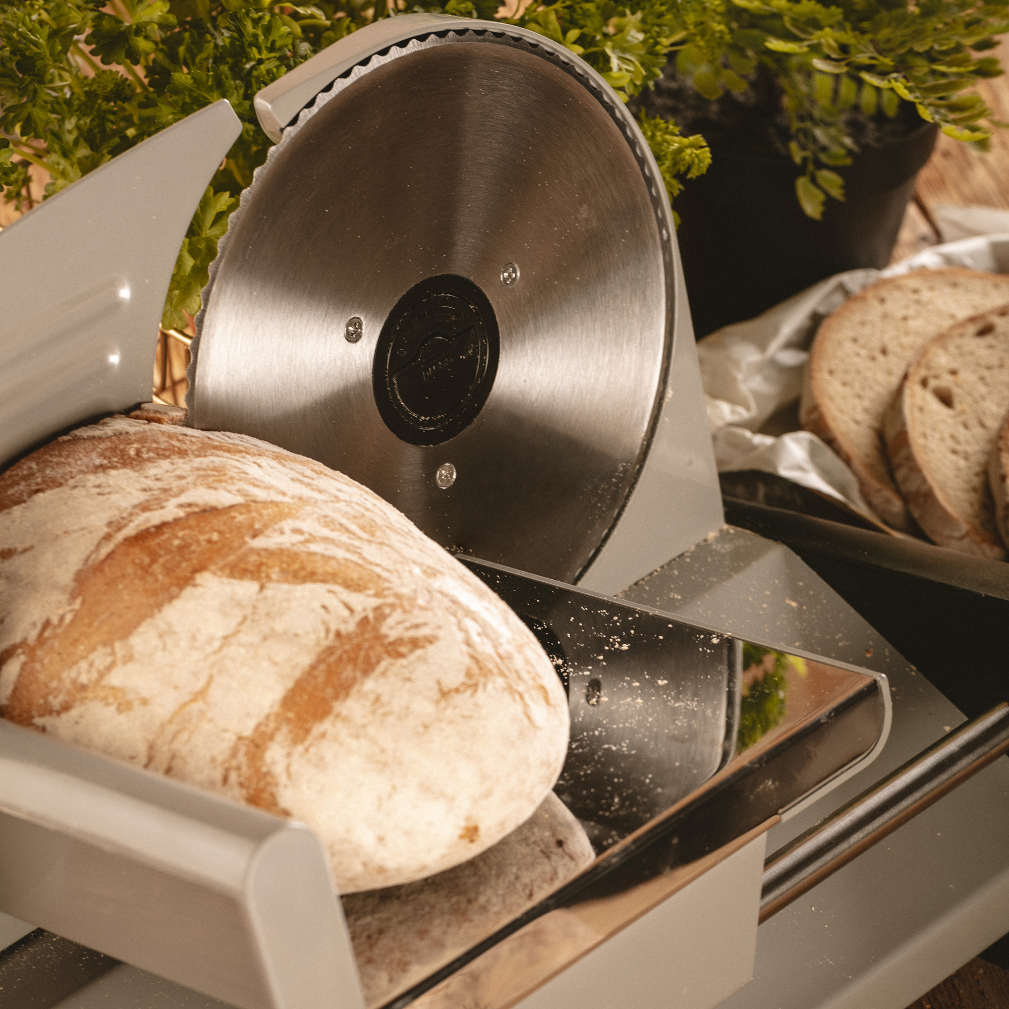 Krojalnica do chleba z bochenkiem chleba z przodu, ostrzem tnącym i zielenią w tle.