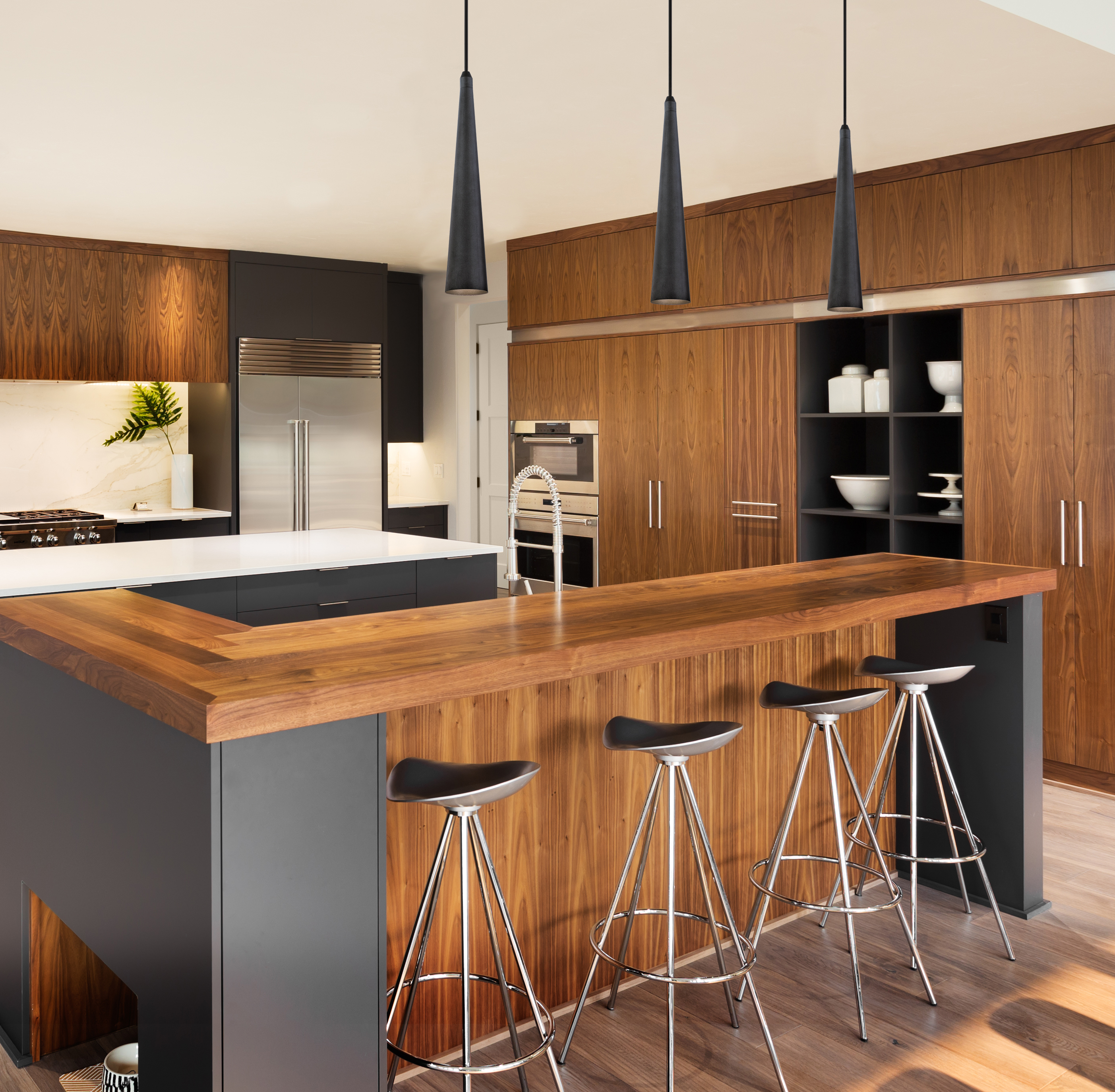 Küche mit Holztisch, schwarzen Hockern und Hängelampen. Holzschränke und ein Kühlschrank aus Edelstahl.