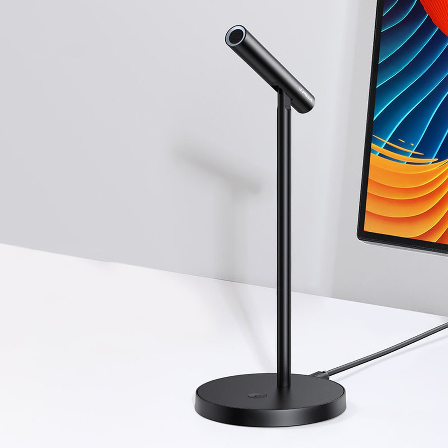 Schwarzes Mikrofon auf einem Ständer, auf einem weißen Schreibtisch vor einem Monitor.