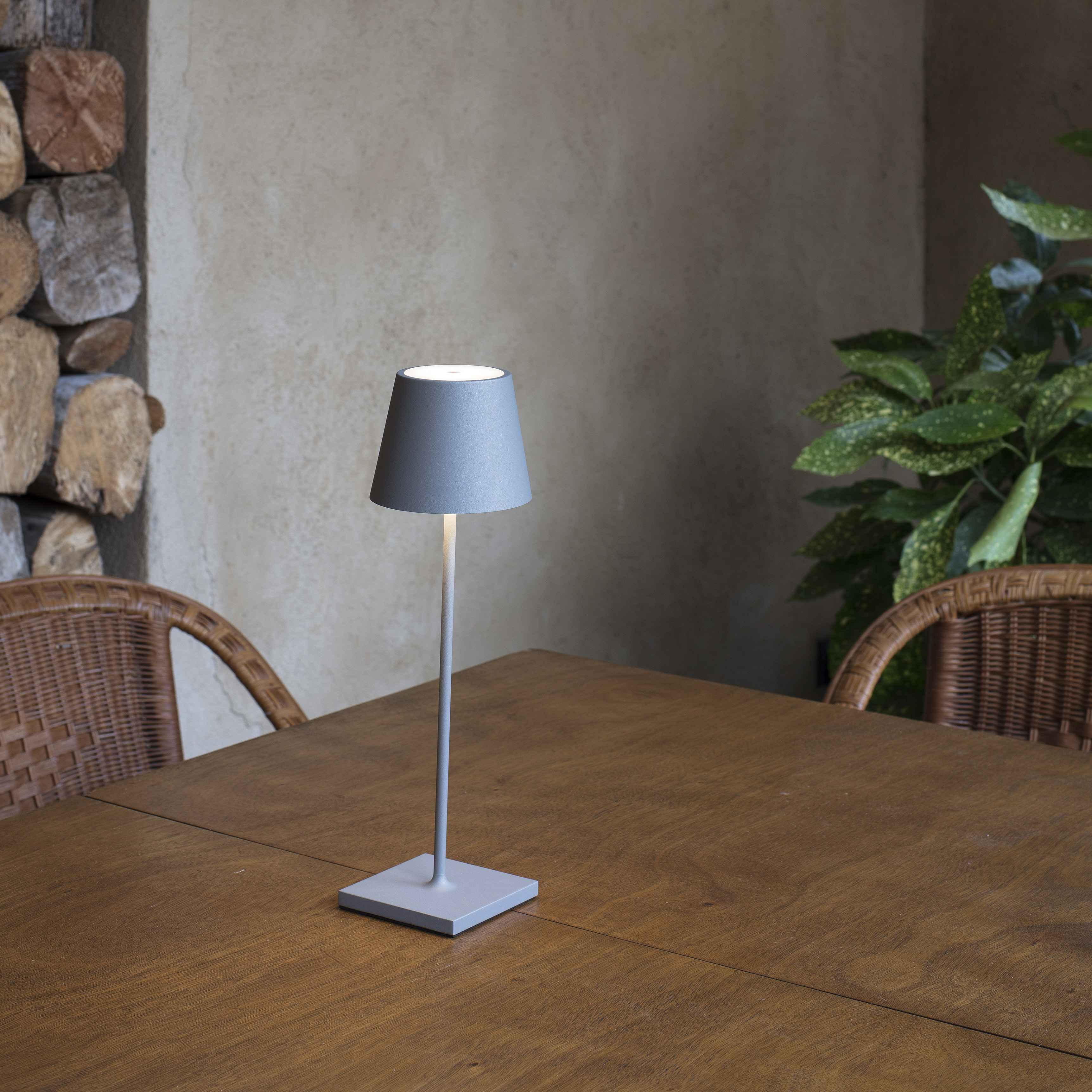 Una lampada grigia su un tavolo di legno, con un muro beige e una sedia di vimini sullo sfondo.