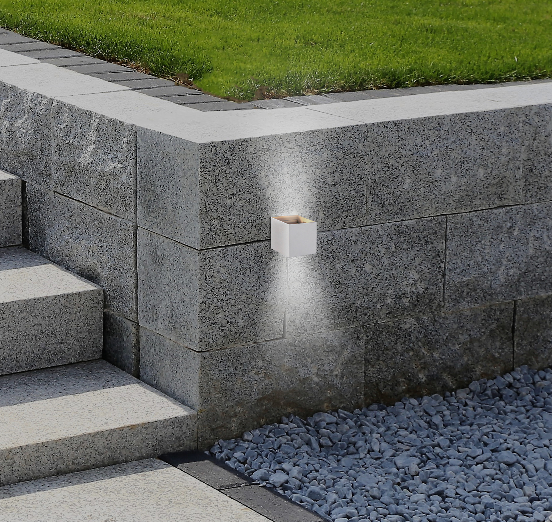 Eine weiße Leuchte an einer grauen Steinmauer. Grünes Gras und Kies im Hintergrund.