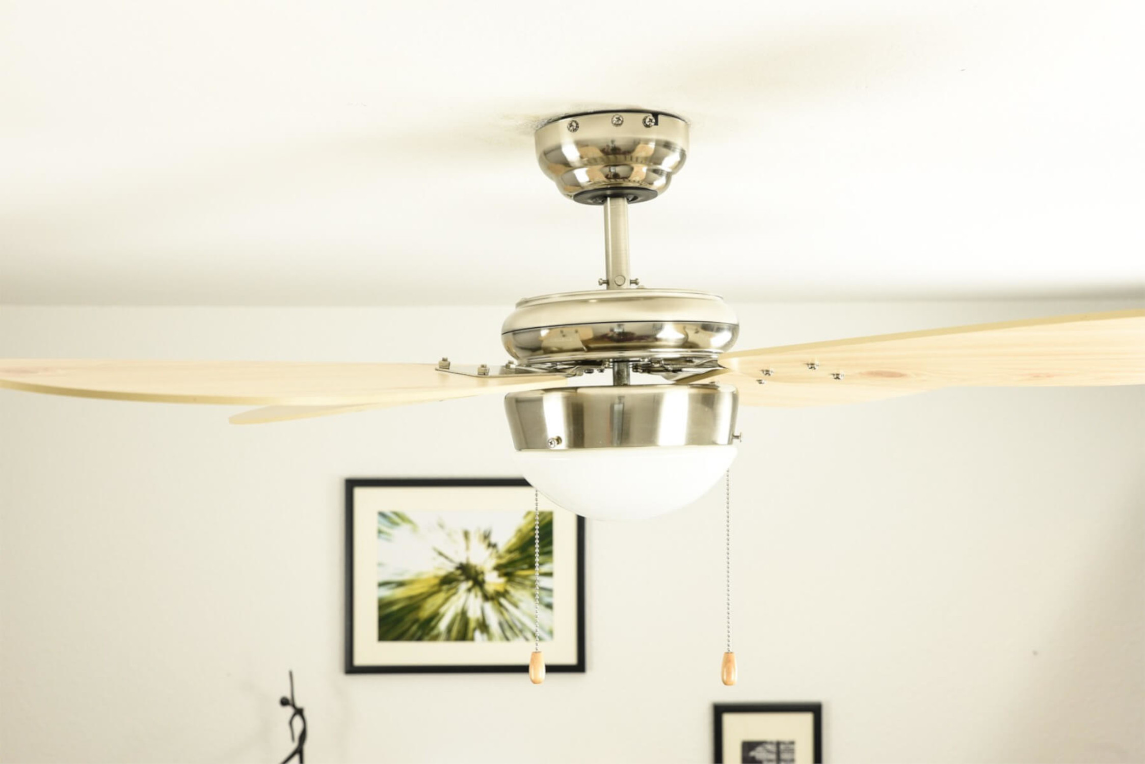 Deckenventilator mit Leuchte, Holzblätter, zwei gerahmte Bilder an der Wand.