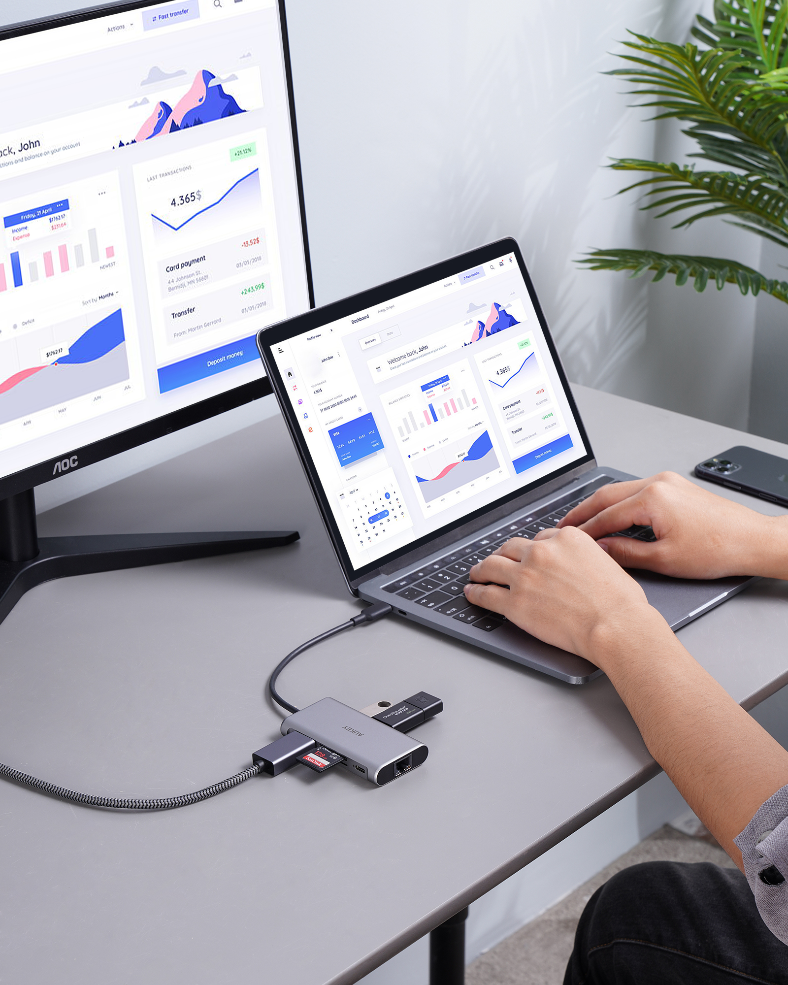 Person nutzt Laptop, verbunden mit Monitor über Hub. Diagramme auf Bildschirmen, Telefon daneben.