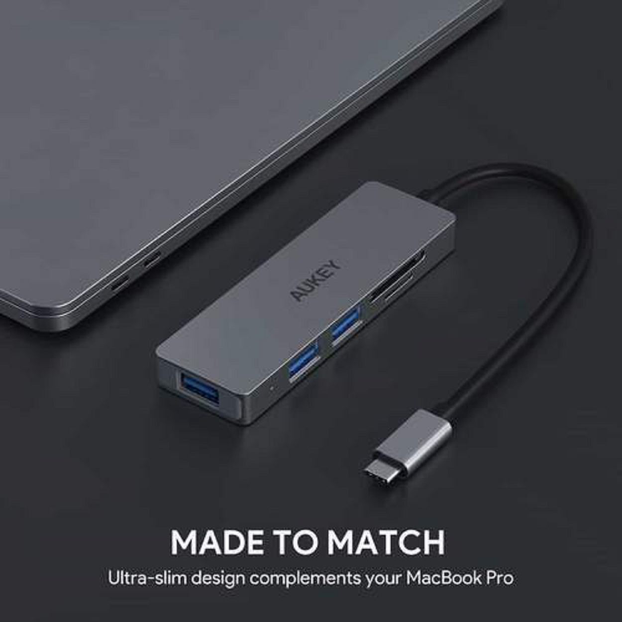 Grauer Hub mit USB-Anschlüssen und einem USB-C-Anschluss, neben einem silbernen Laptop. Dunkelgrauer Hintergrund.