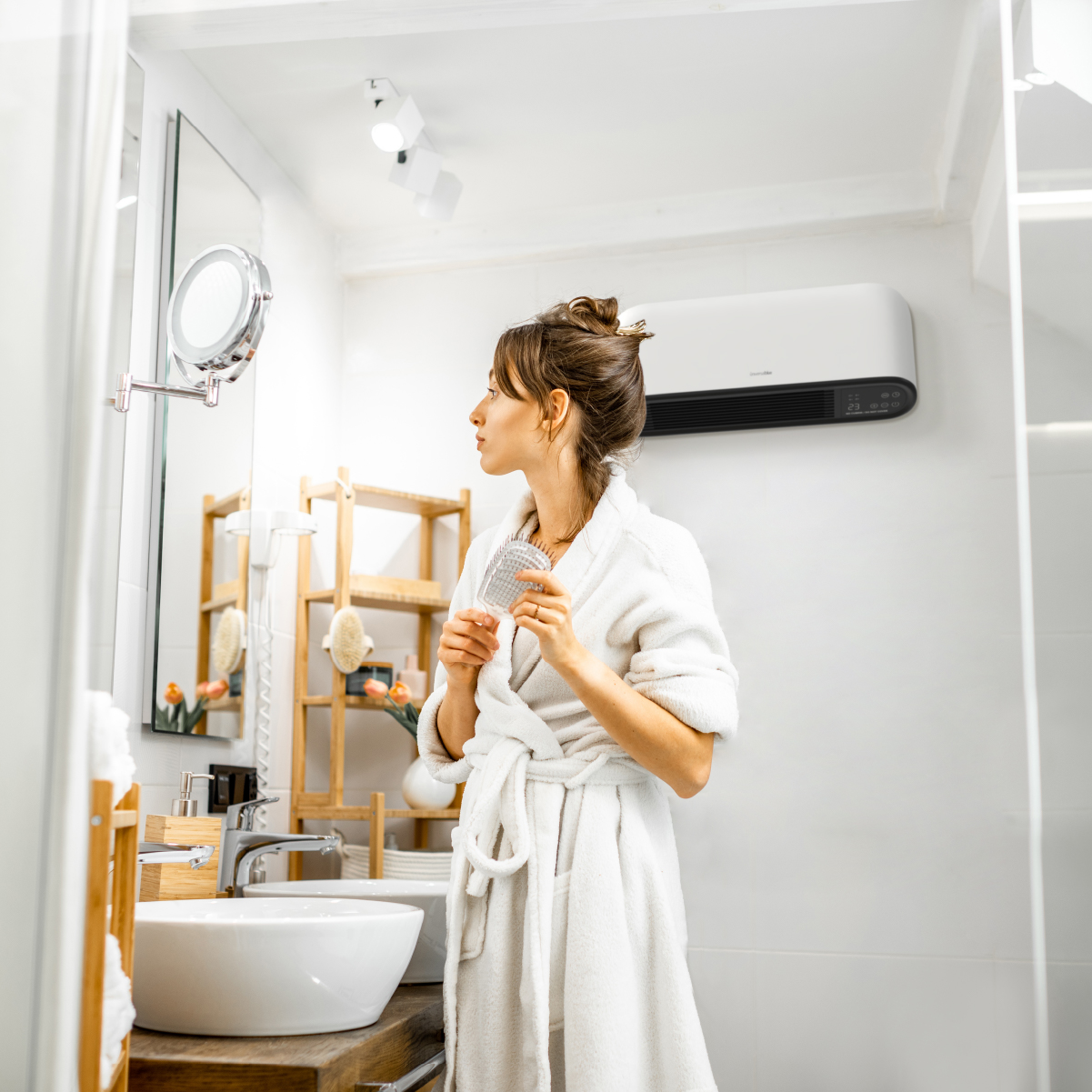 Kobieta w białym szlafroku w łazience, patrząca w lustro z szczotką do włosów.