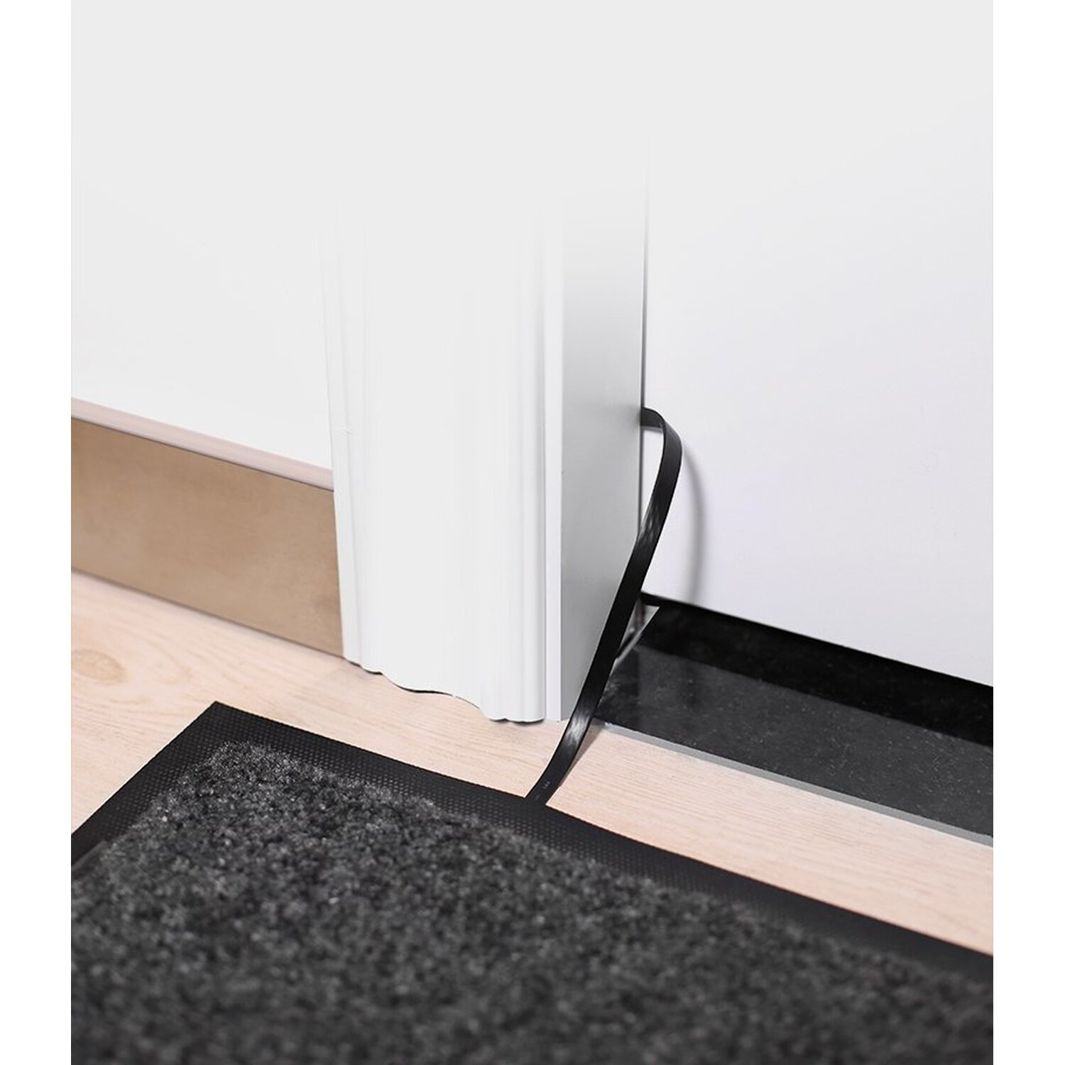 Czarna i szara wycieraczka umieszczona na jasnej drewnianej podłodze obok białych drzwi. Mata jest teksturowana.