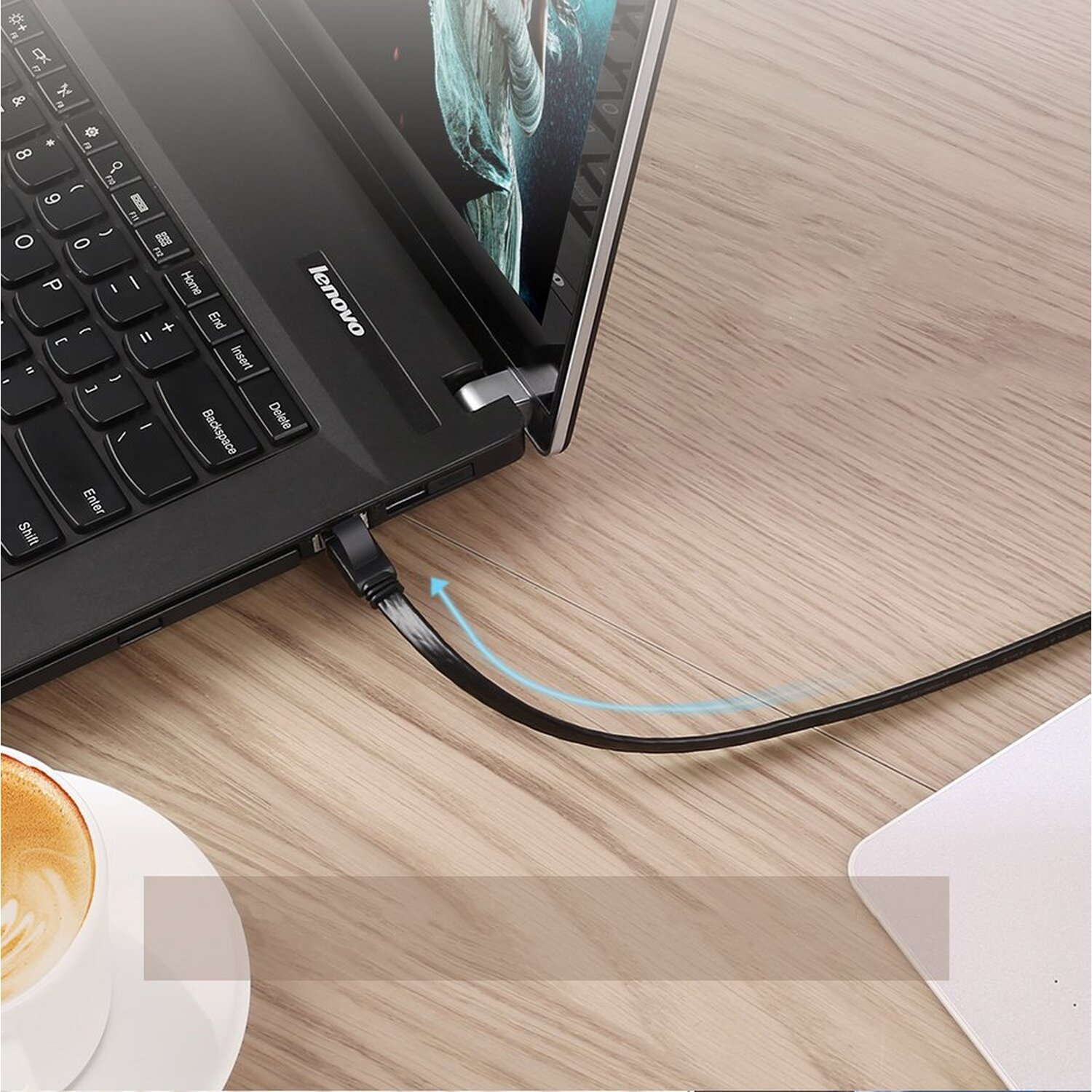 Laptop Lenovo nero con cavo nero collegato su tavolo in legno, con tazza di caffè.