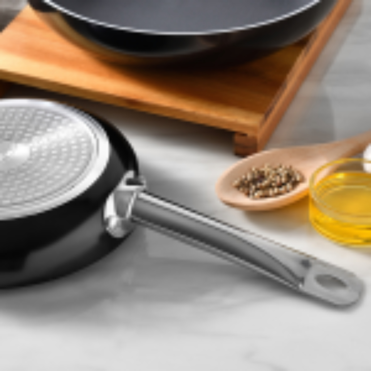 Primer plano de una sartén negra con mango plateado. Se ven una tabla de madera, aceite de oliva y especias.