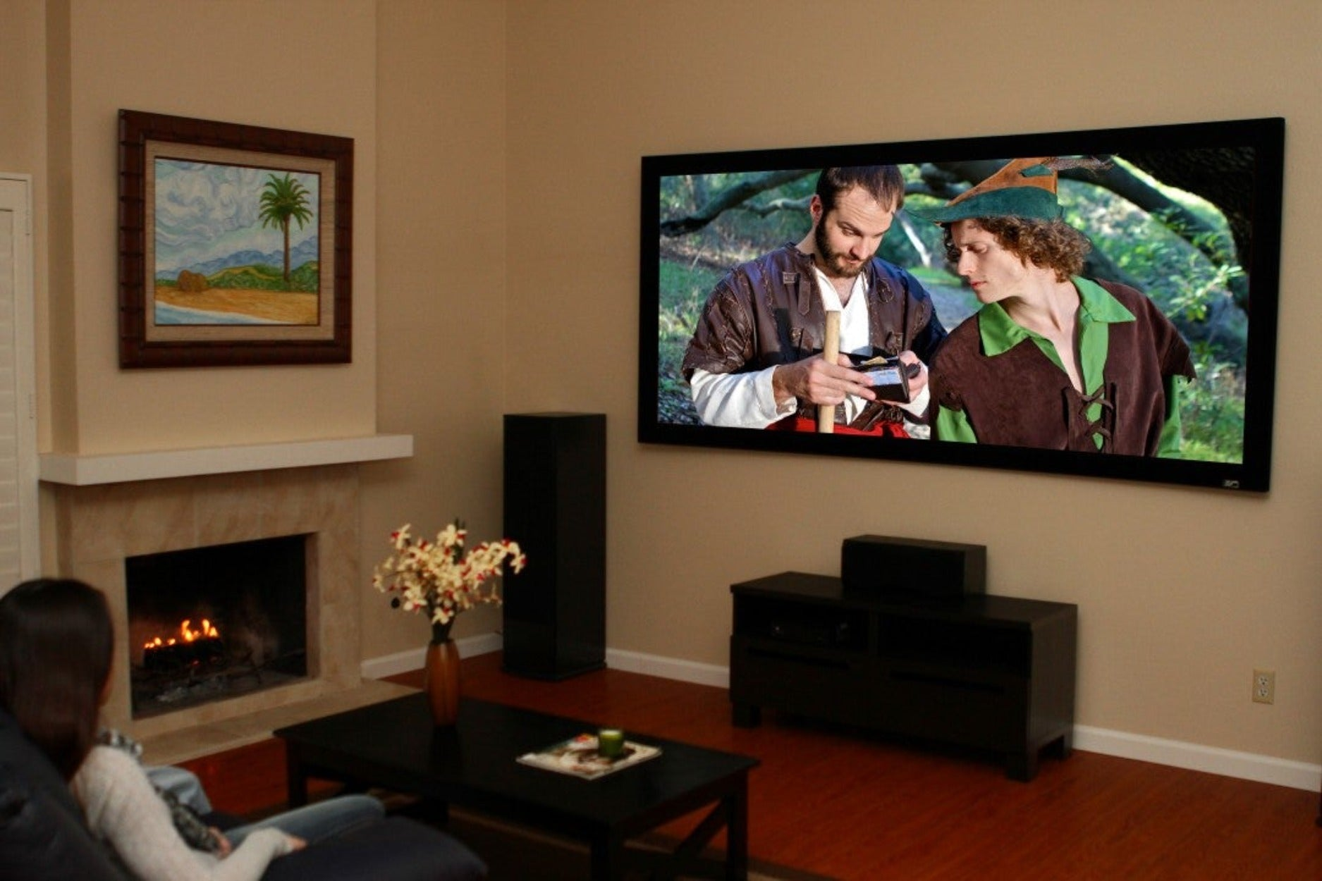 Soggiorno con grande TV a parete, persone che guardano, camino e un quadro incorniciato.