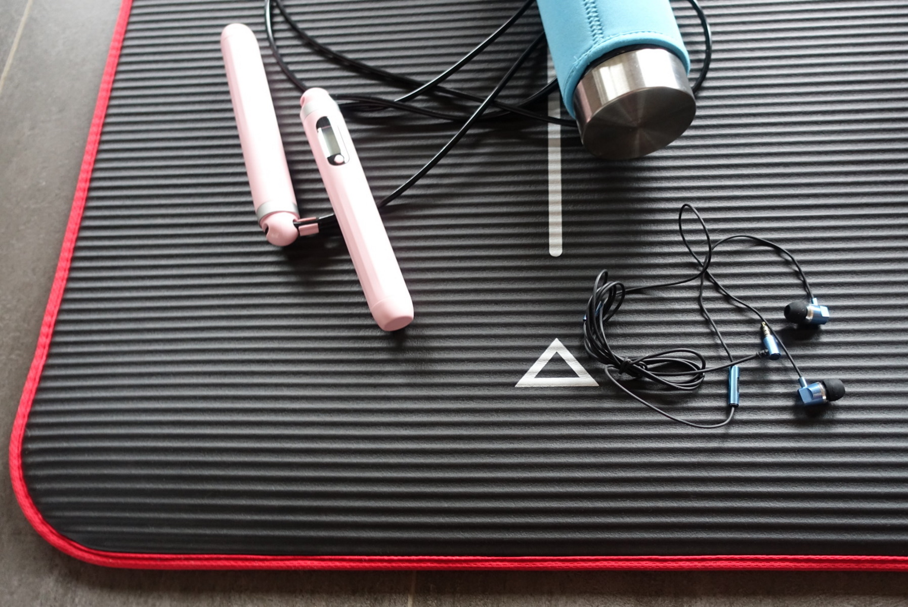 Workout-Matte mit rosa Springseil, Kopfhörern und Wasserflasche, alles auf schwarzer Oberfläche.