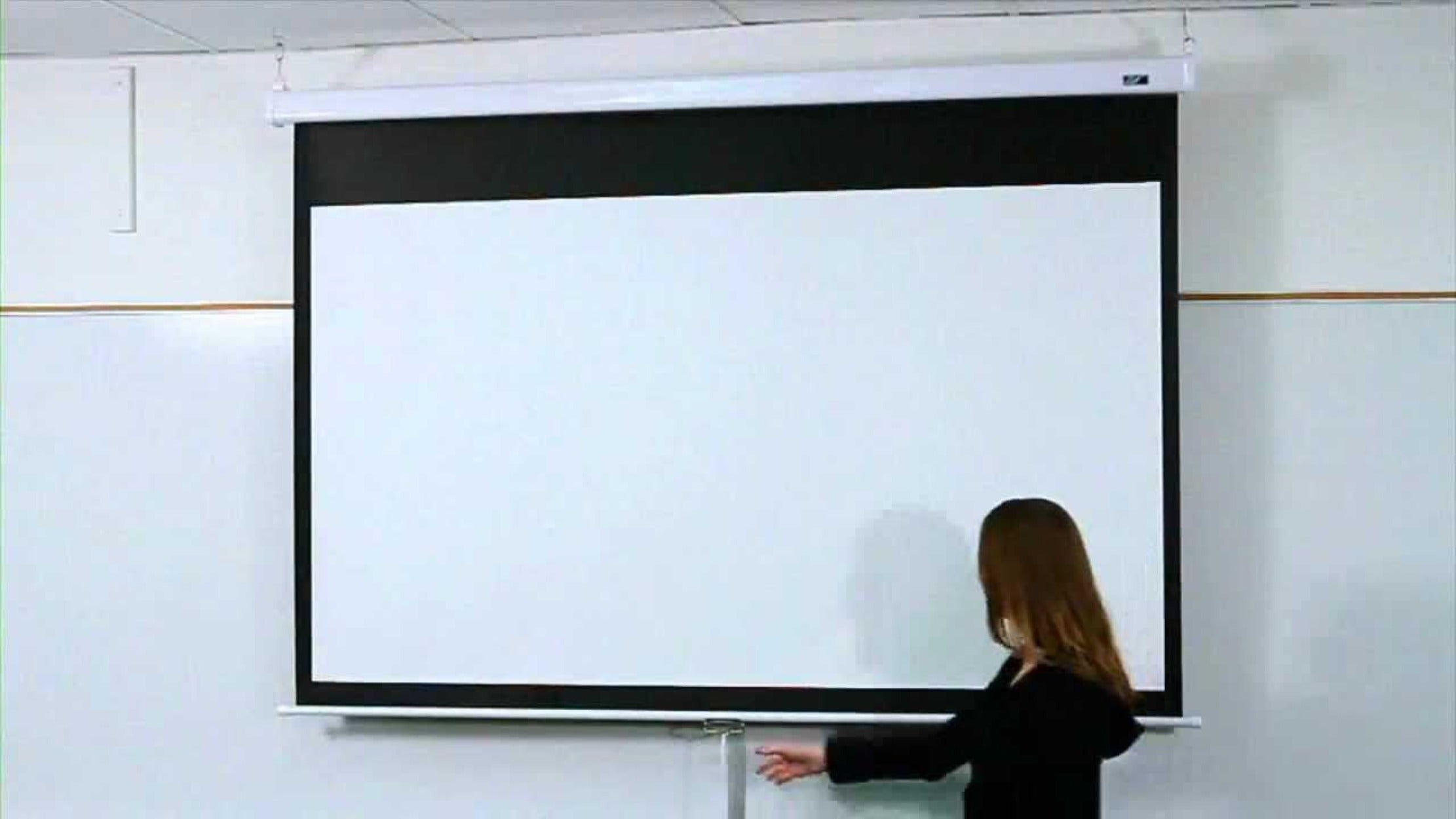 Eine Frau justiert eine weiße Projektionsleinwand, die an einer weißen Wand montiert ist.