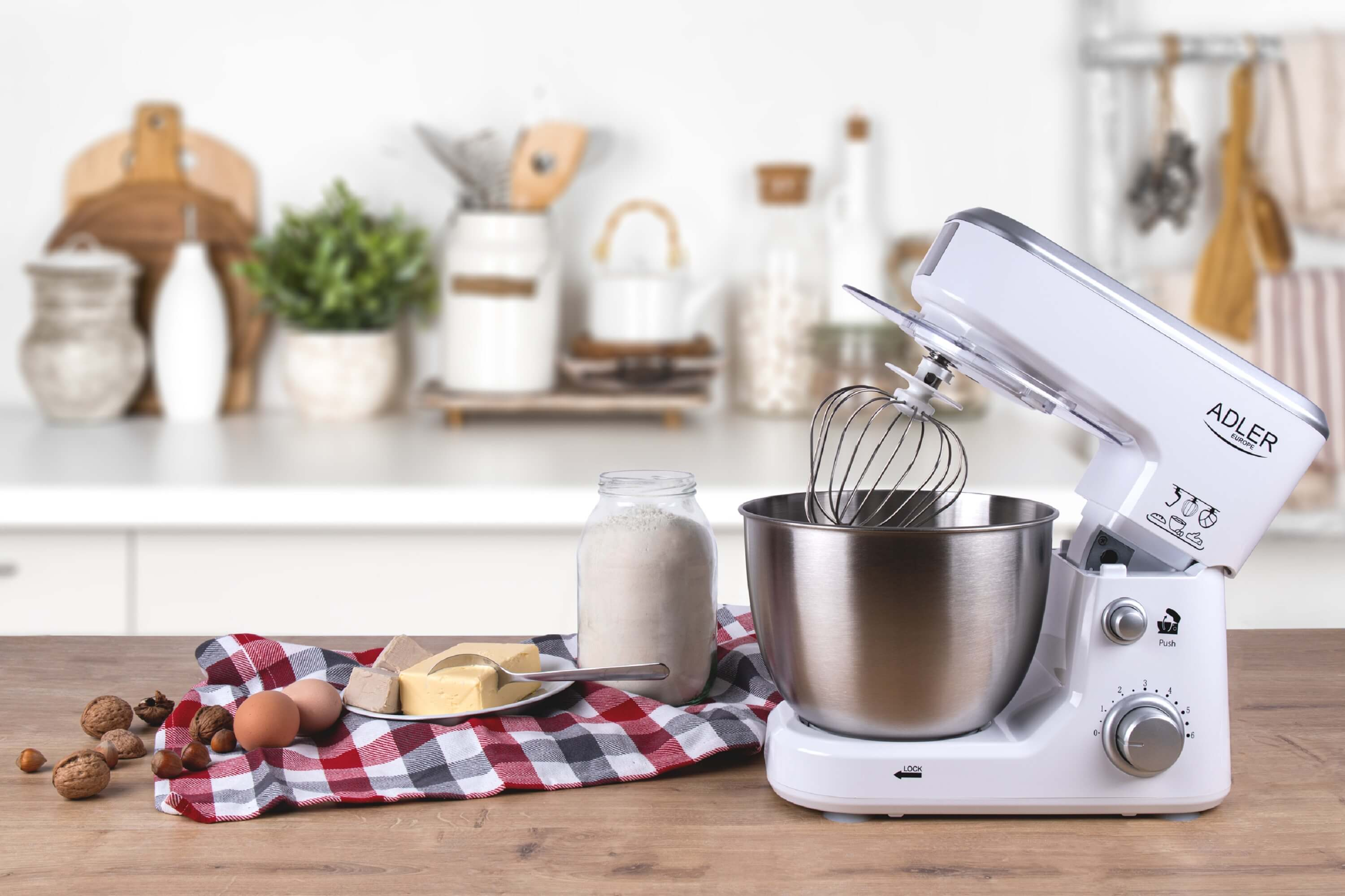 Biały mikser stojący z akcesoriami na drewnianym stole. Widoczne składniki, takie jak jajka, mąka i masło.