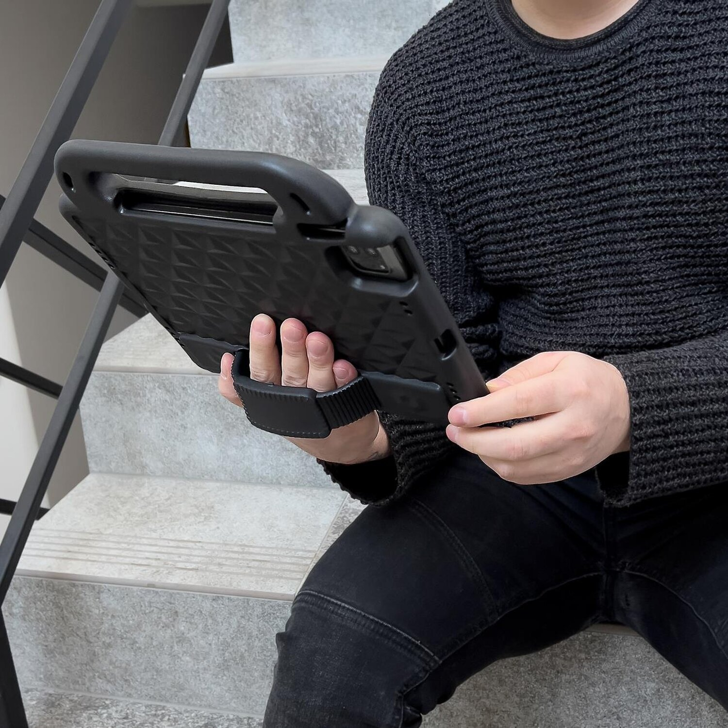 Person hält ein schwarzes Tablet mit Griff, sitzt auf Treppen. Diamantmuster sichtbar.