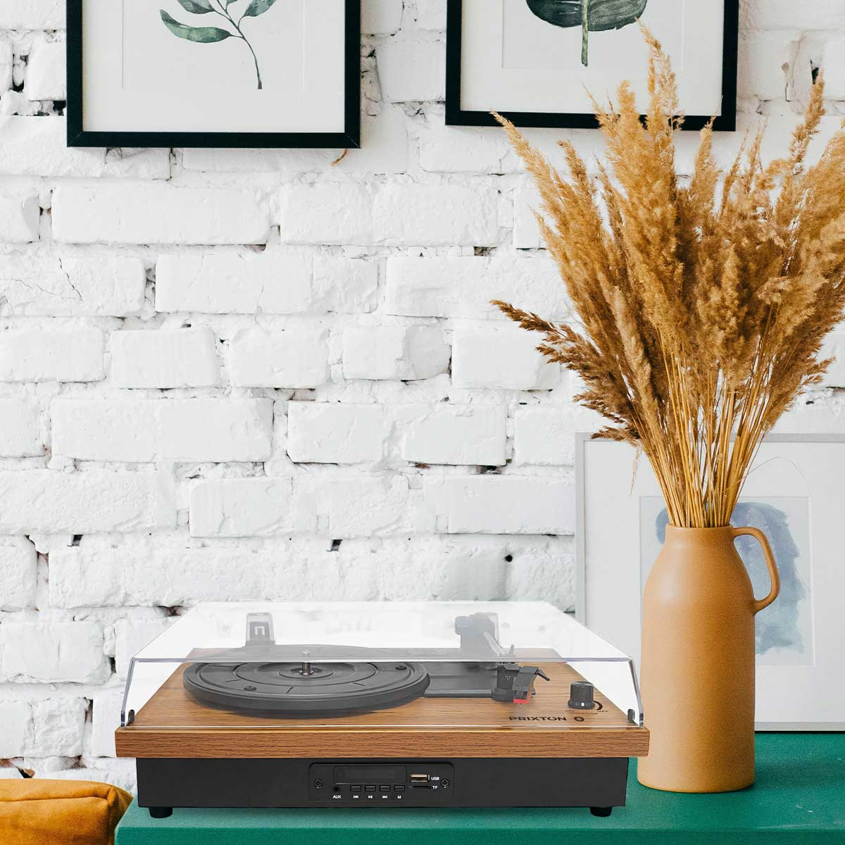 Drewniany gramofon z przezroczystą pokrywą na zielonej powierzchni, z roślinami i obrazami w tle.