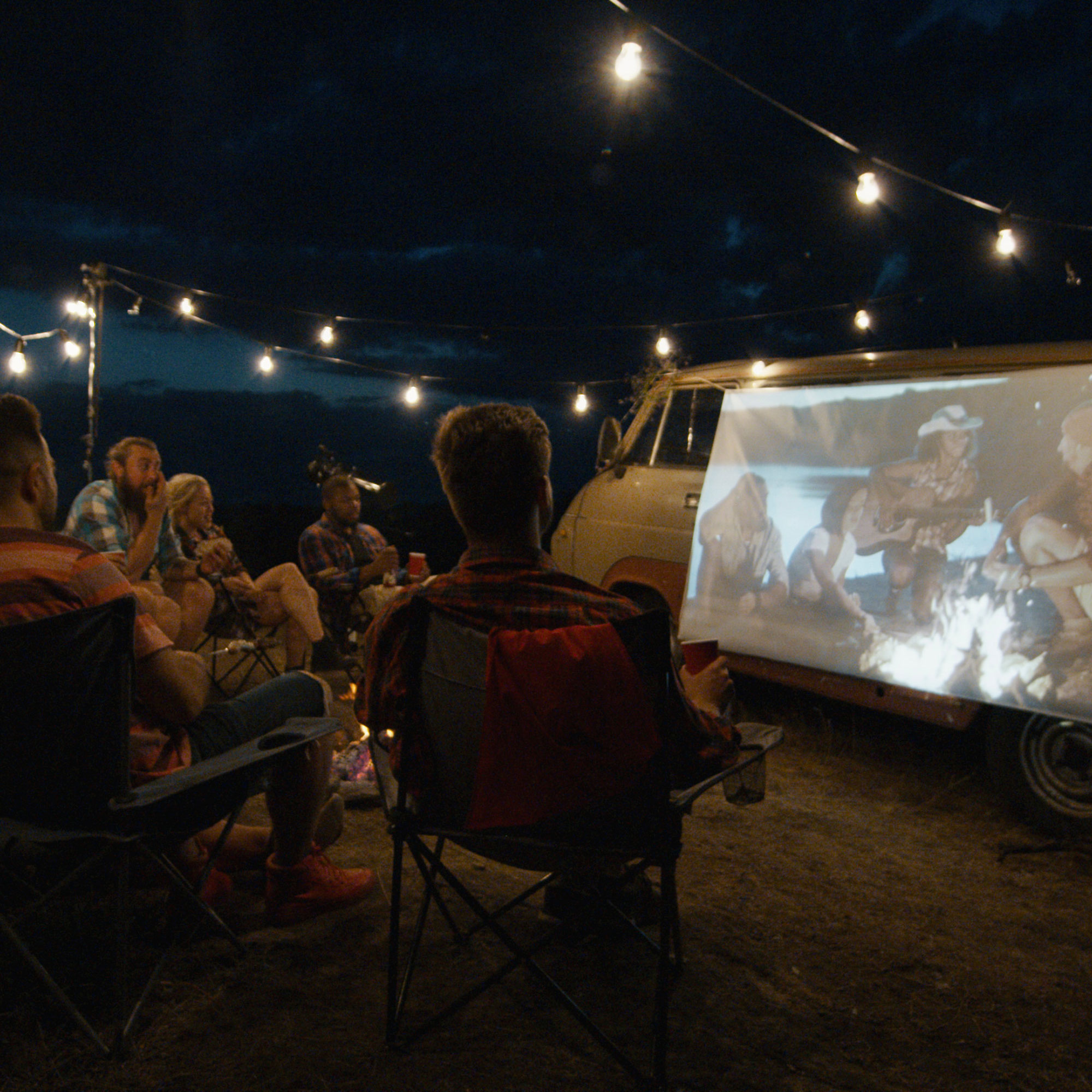 Groupe de personnes regardant un film projeté sur une camionnette. Nuit sombre avec guirlandes lumineuses.