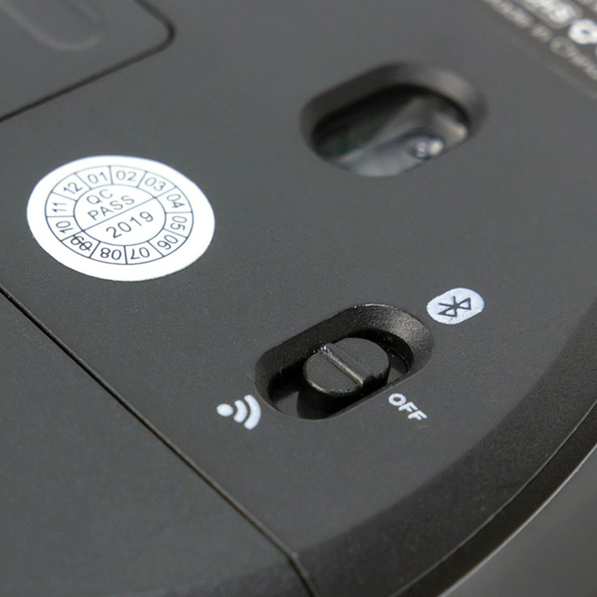 Een close-up van de onderkant van een zwarte muis met een Bluetooth-symbool en een uit-schakelaar. Het heeft een QC Pass 2019-sticker.