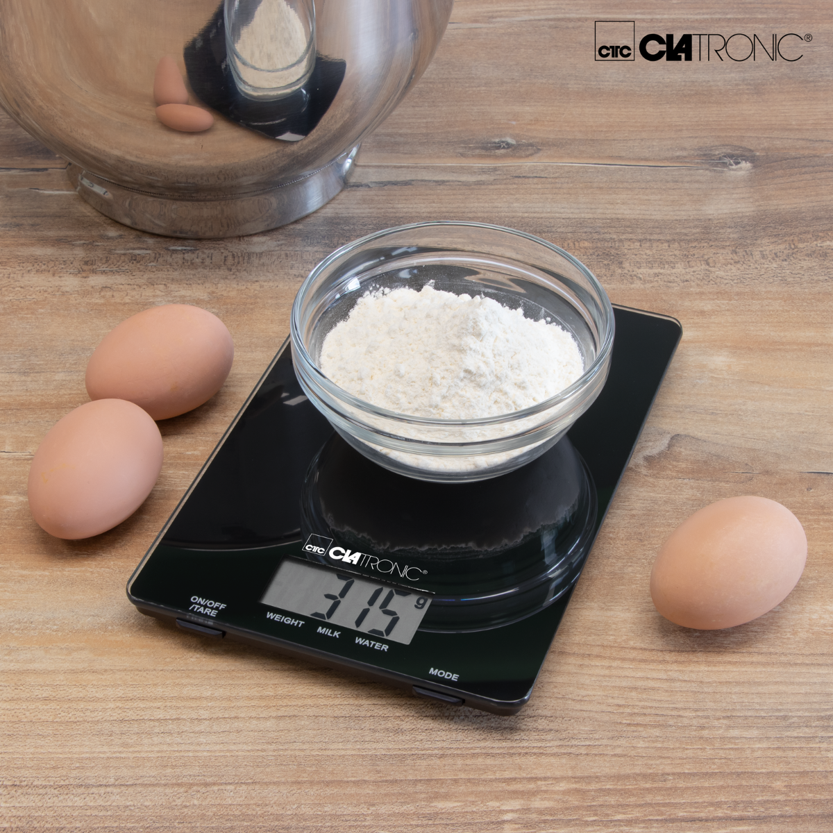 Báscula digital negra con ingredientes, incluyendo huevos y harina en un cuenco. La báscula muestra un peso.