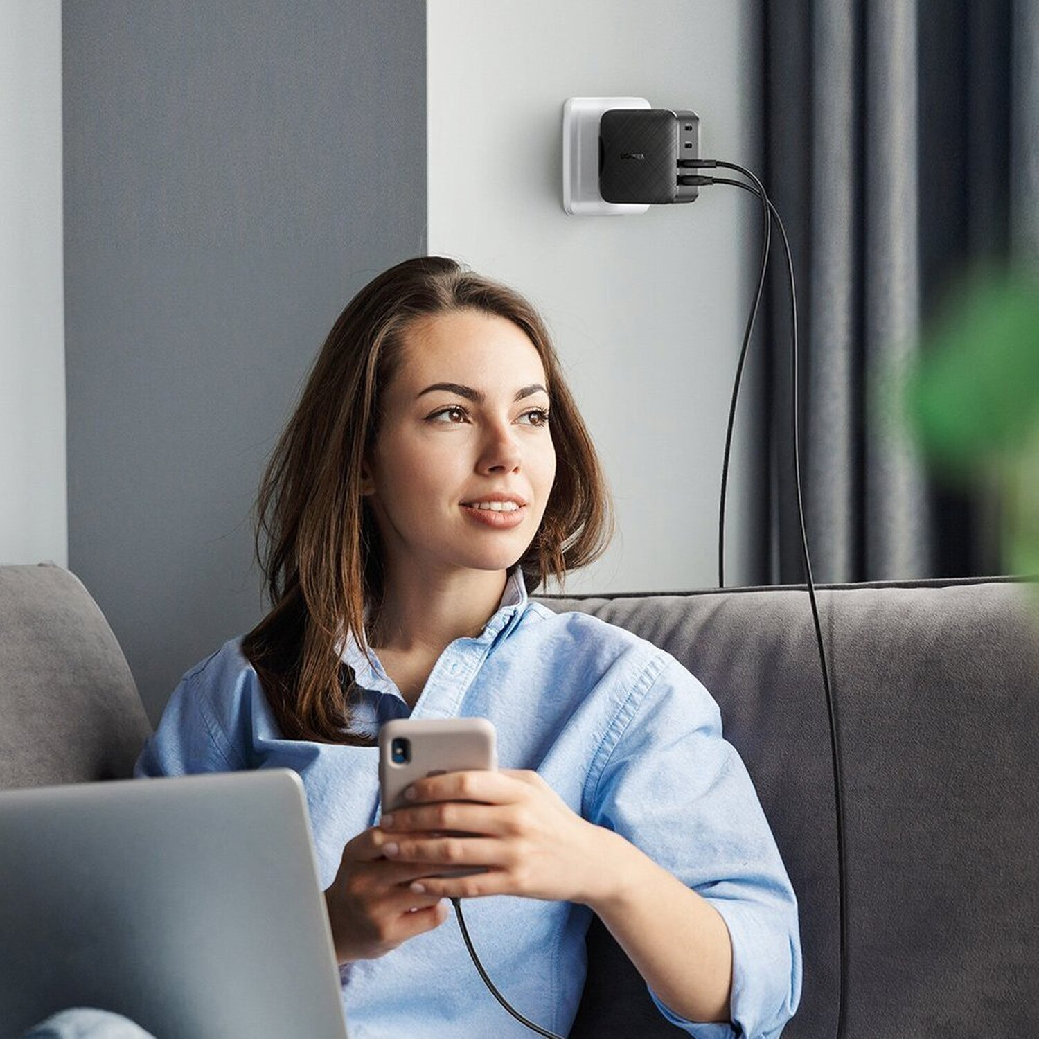 Frau auf Sofa mit Telefon, Laptop und Wandladegerät. Lächelt und schaut weg. Graues Sofa, weiße Wand.