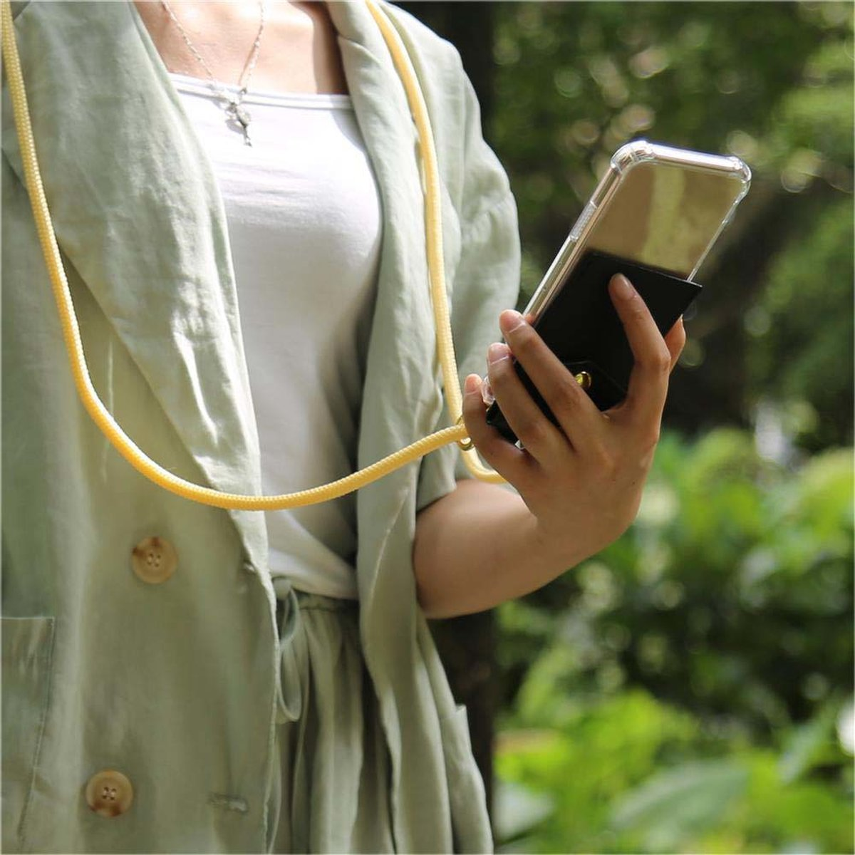 Una donna tiene in mano un telefono con un cordino giallo al collo. Indossa una giacca verde e un top bianco. Fogliame verde sullo sfondo.
