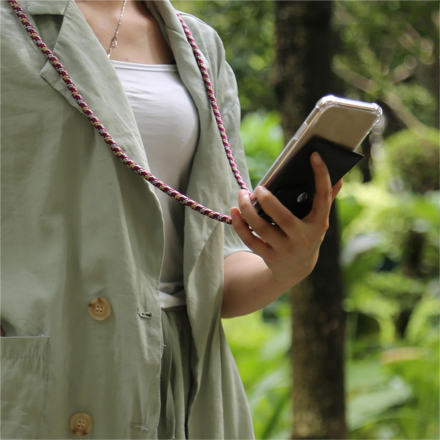 Una persona tiene in mano un telefono in una custodia nera con un cordino rosso/bianco. Lo sfondo è un parco verde.