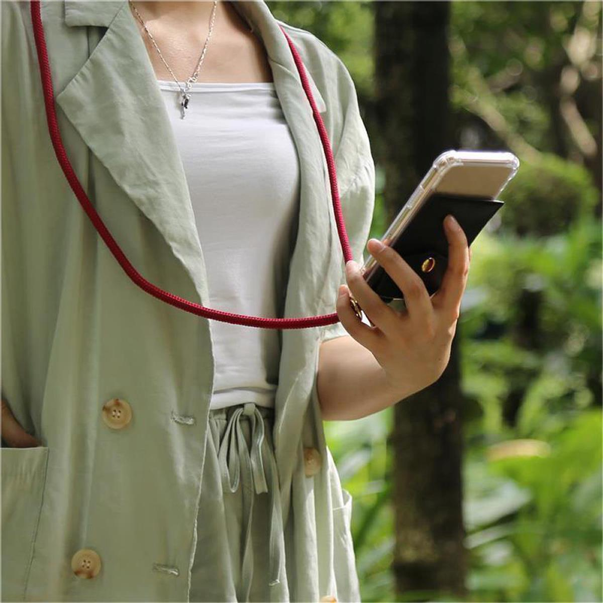 Una donna che tiene un telefono in una custodia con un cordino rosso al collo in un ambiente verde.