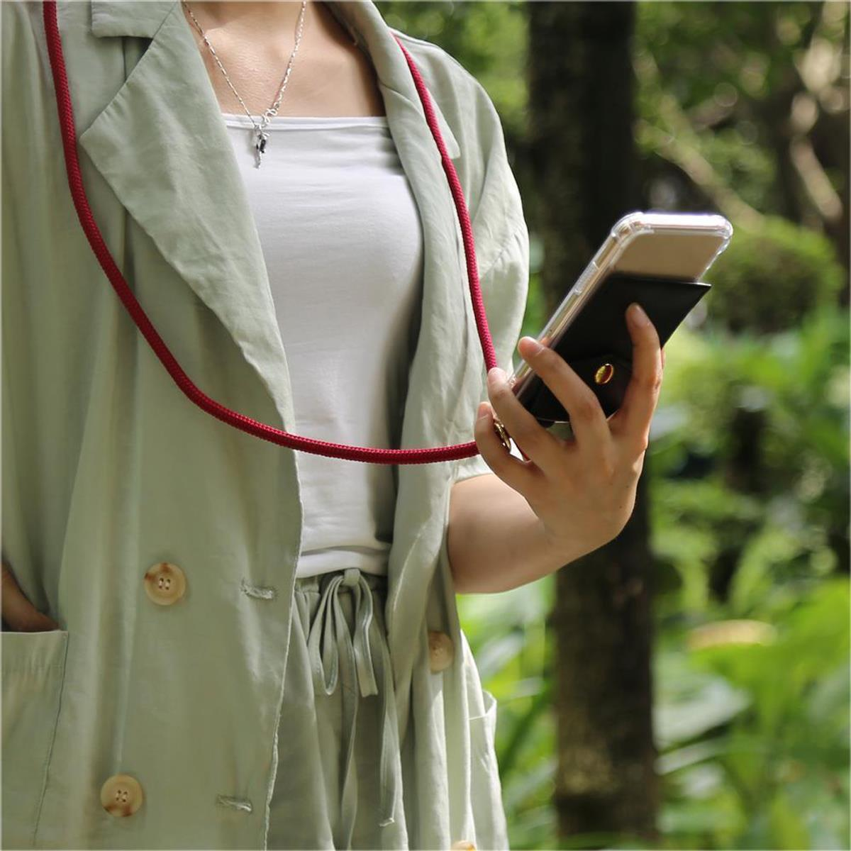 Persona che tiene un telefono con custodia, indossa collana e giacca, in un giardino.