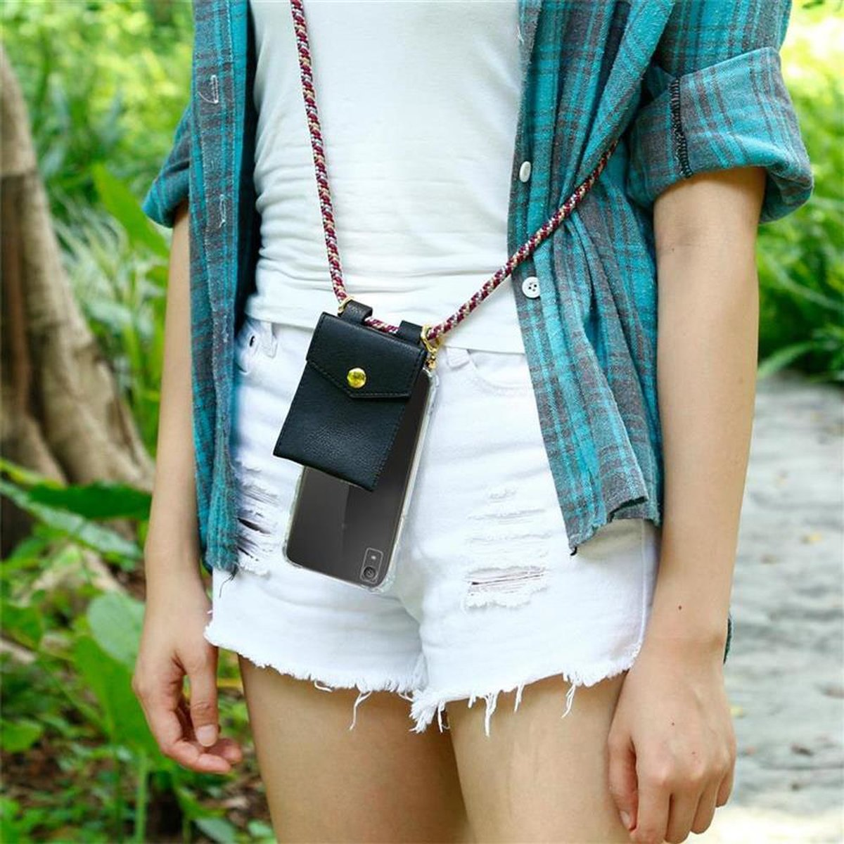 Frau in weißem Top, Shorts, blauem karierten Hemd, hält Telefon in Hülle mit Riemen. Hintergrund: Bäume und ein Weg.