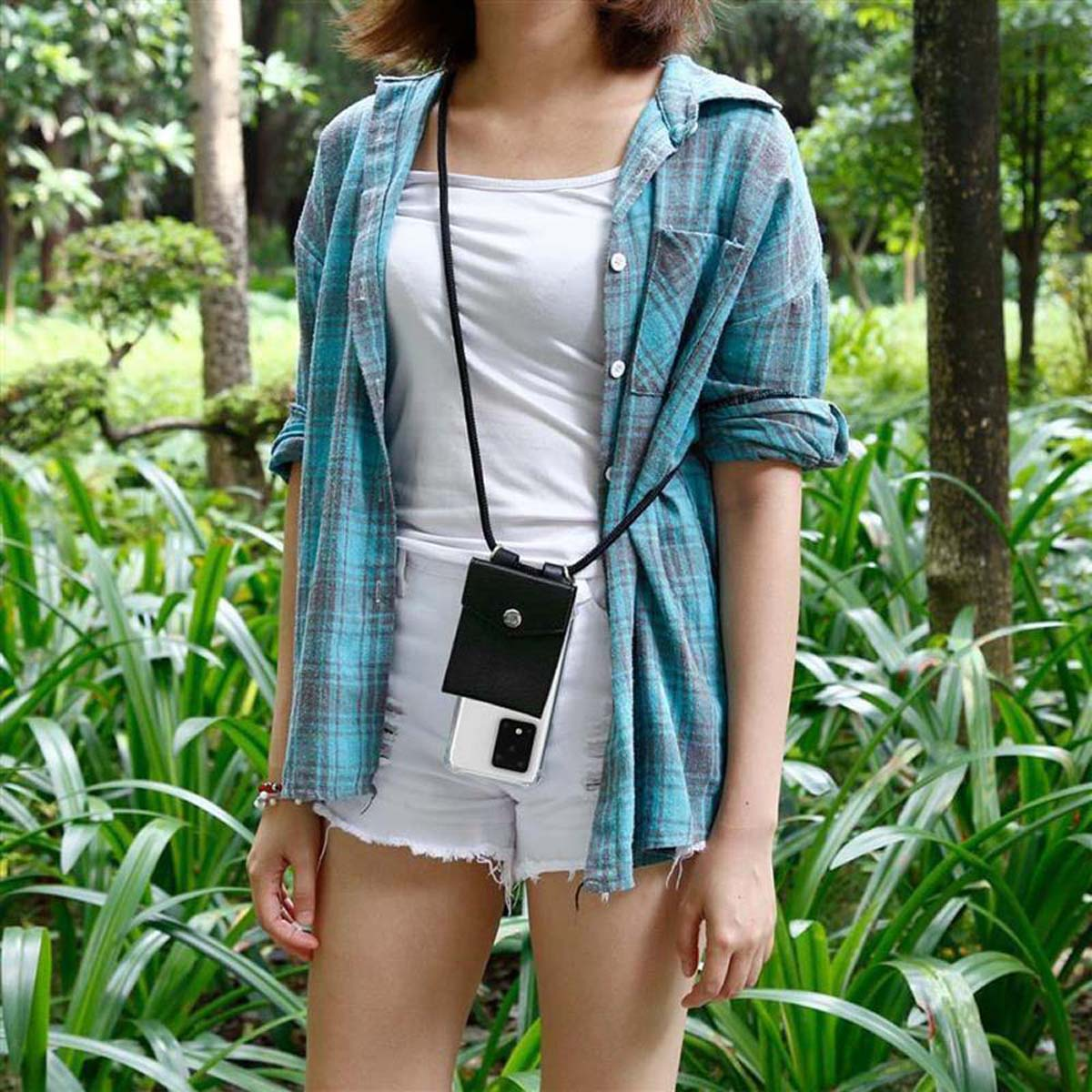 Donna con pantaloncini bianchi, camicia a quadri e una custodia per telefono con cinturino nero in un parco.