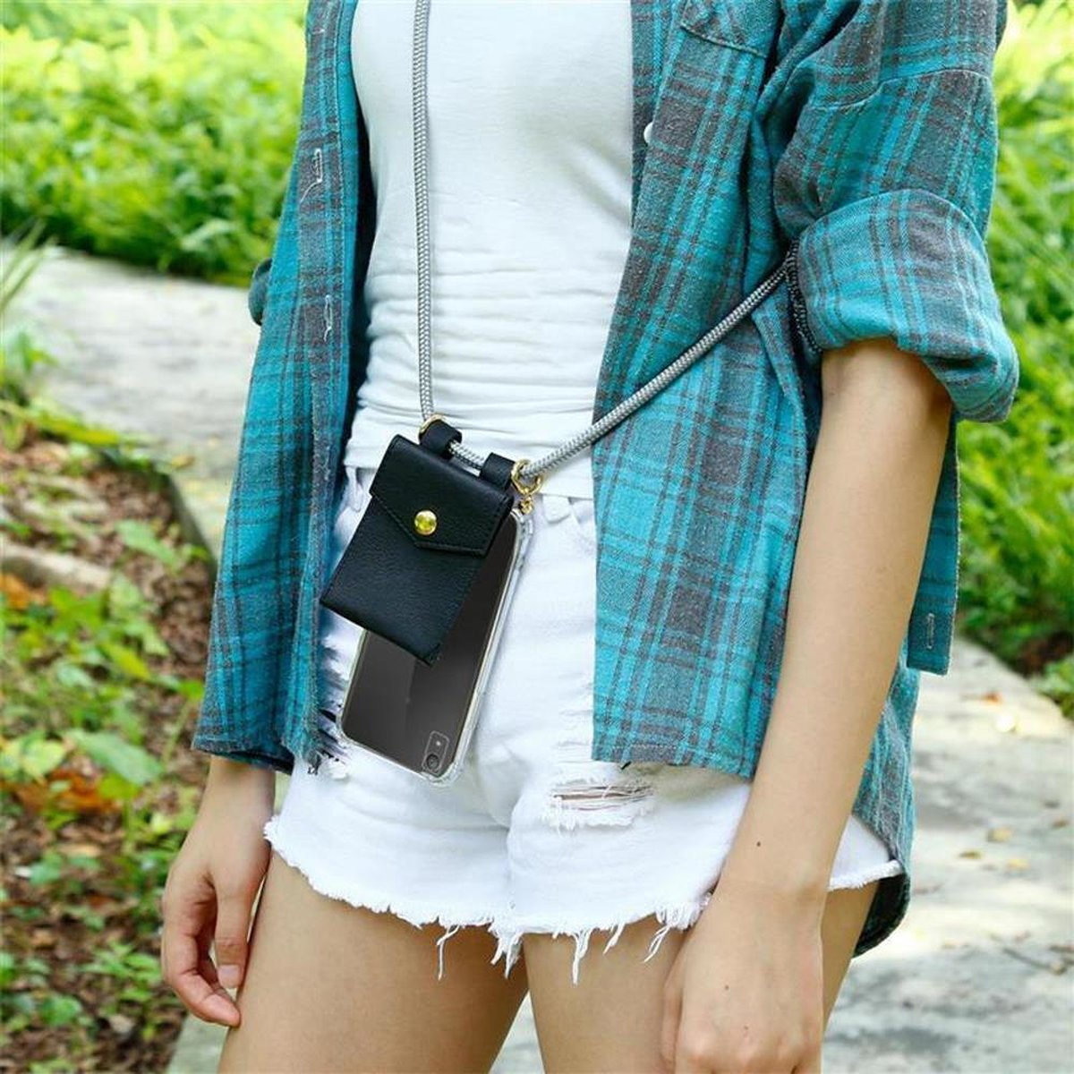 Frau mit blauem karierten Hemd, weißen Shorts, mit einer Handyhülle mit Riemen, in einer Parkumgebung.