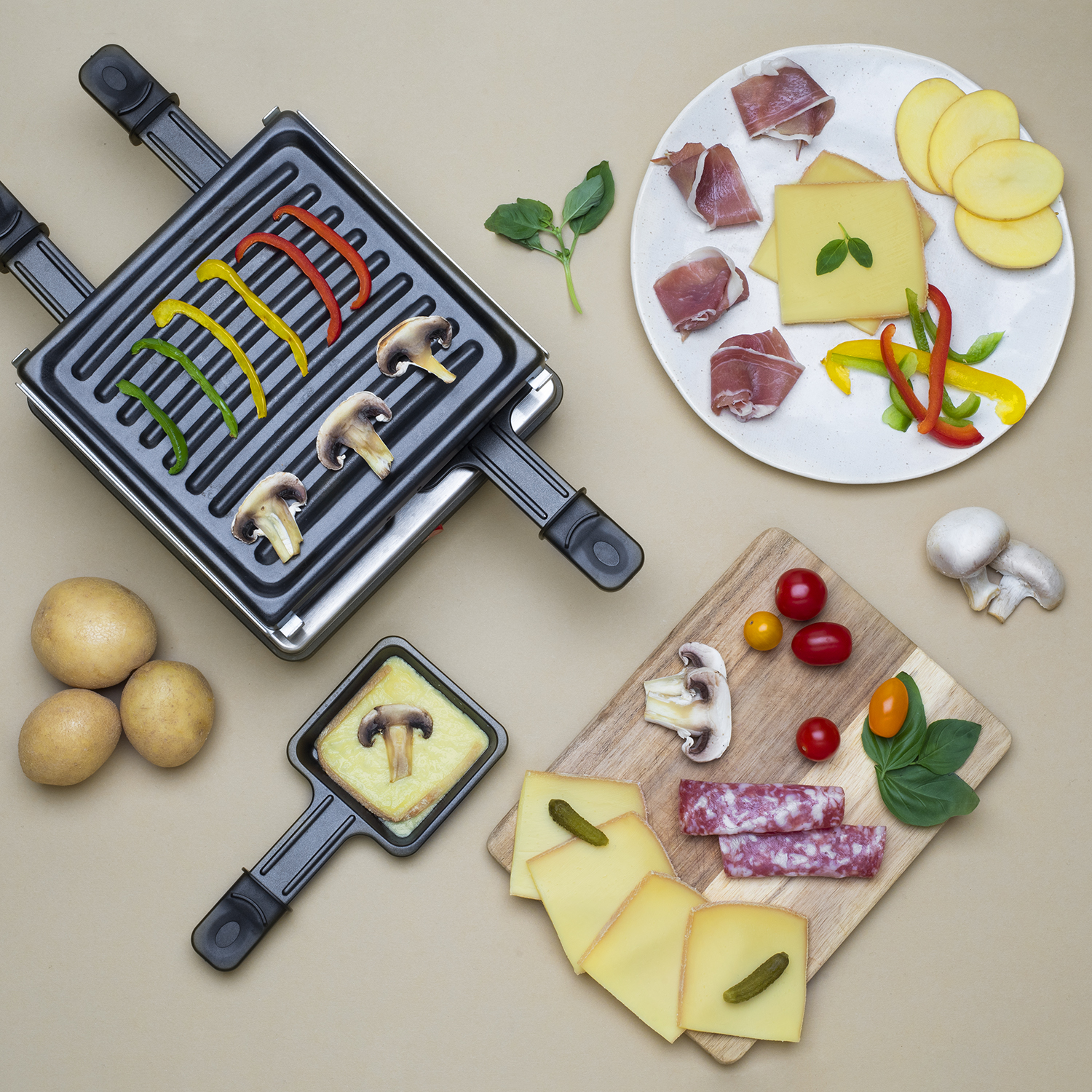 Raclette-Grill mit Gemüse, Käse, Fleisch und Kartoffeln auf beiger Oberfläche.