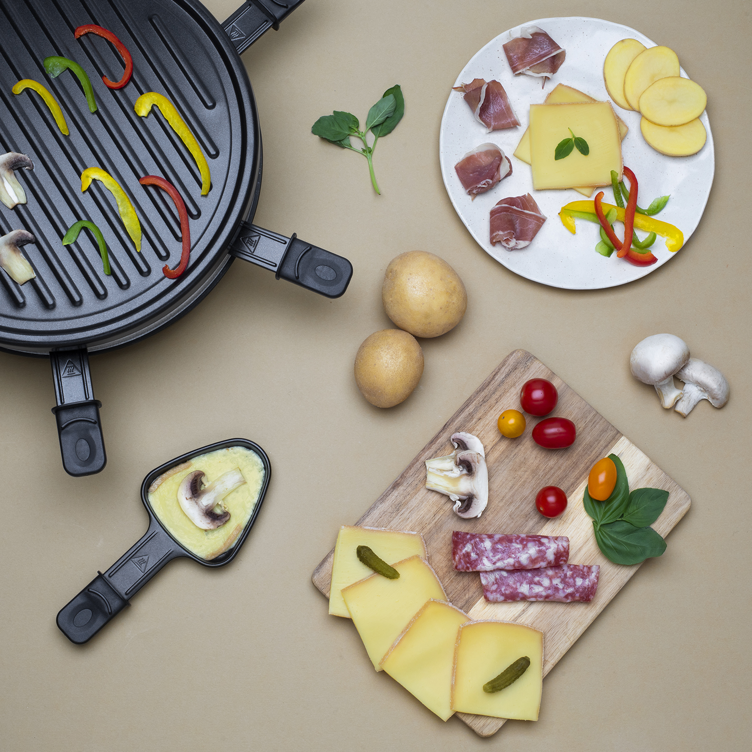 Raclette-Grill mit Gemüse, Käse, Kartoffeln und Salami, auf beiger Oberfläche.