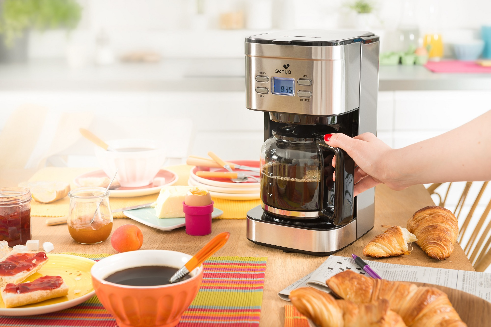 Scena della colazione con macchina del caffè, croissant, marmellata, uova e caffè. Mano di donna.